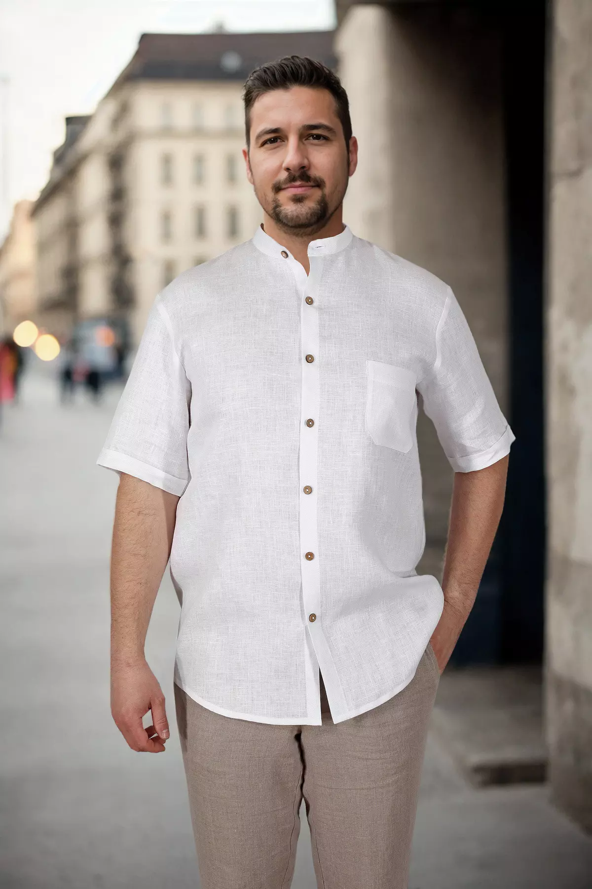 Mężczyzna stoi prosto na tle miejskiej architektury. Prezentuje białą koszulę Tom ze stójką. Koszula jest wypuszczona na wierzch beżowych spodni. Kontrastowe guziki i kieszonka są wyraźnymi detalami.