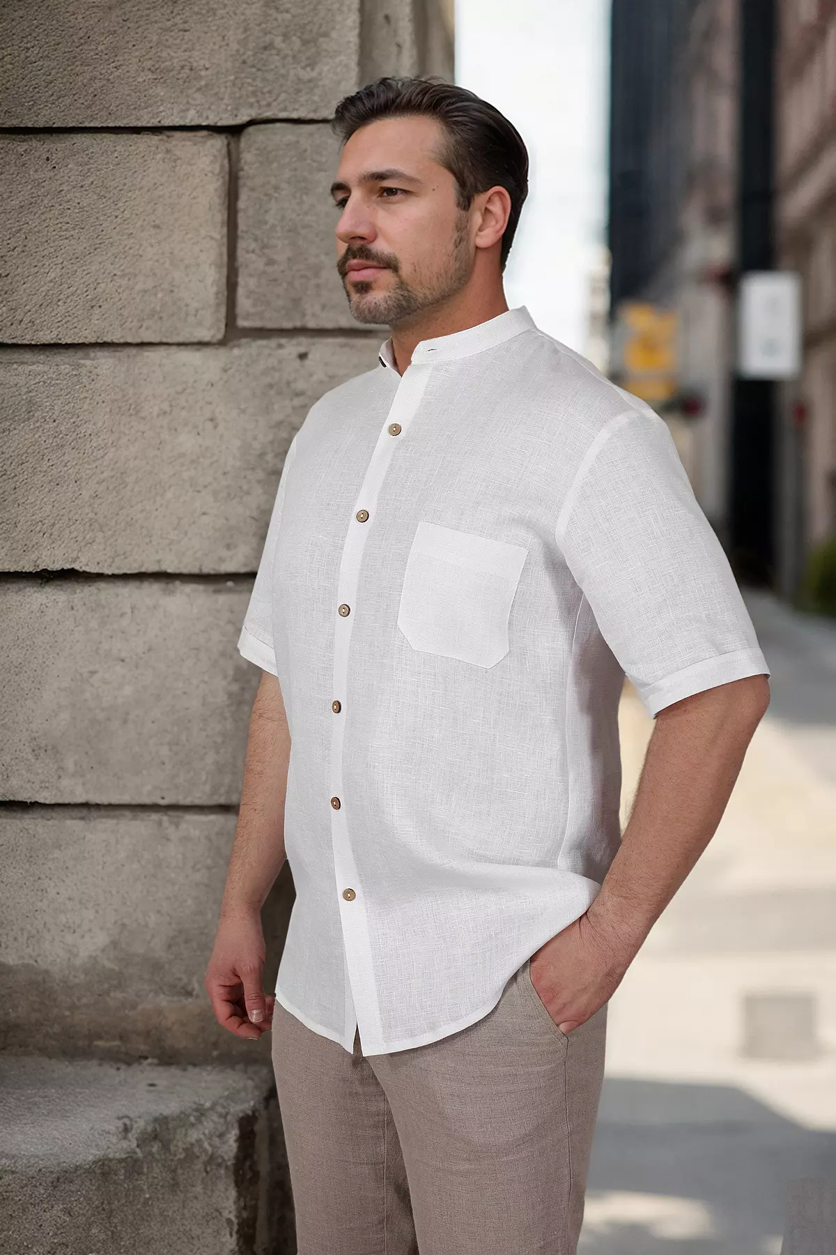 Mężczyzna stoi w półprofilu. Ma na sobie białą koszulę lnianą Tom ze stójką i krótkim rękawem. Koszula jest zapinana na kontrastowe, brązowe guziki. Widać kieszonkę na piersi. Mężczyzna trzyma rękę w kieszeni beżowych spodni.