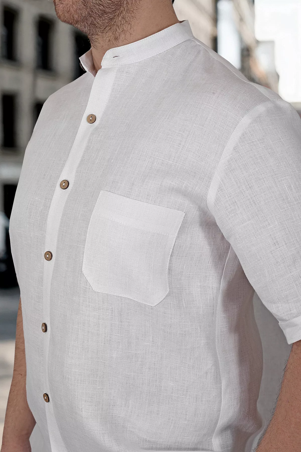 Zbliżenie na górną część koszuli. Widać precyzyjnie wykończoną stójkę oraz naszytą kieszonkę na piersi. Zdjęcie eksponuje naturalną fakturę białej tkaniny lnianej i kokosowe guziki.