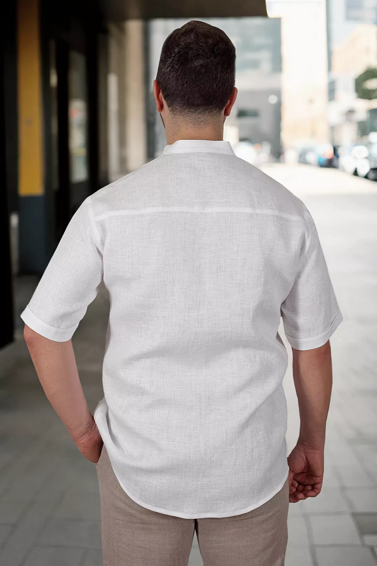 Widok pleców mężczyzny stojącego na chodniku. Koszula Tom ma prosty tył z karczkiem, co zapewnia wygodę. Krótkie rękawy i luźny fason są idealne na ciepłe dni. Biały len wygląda świeżo i naturalnie.