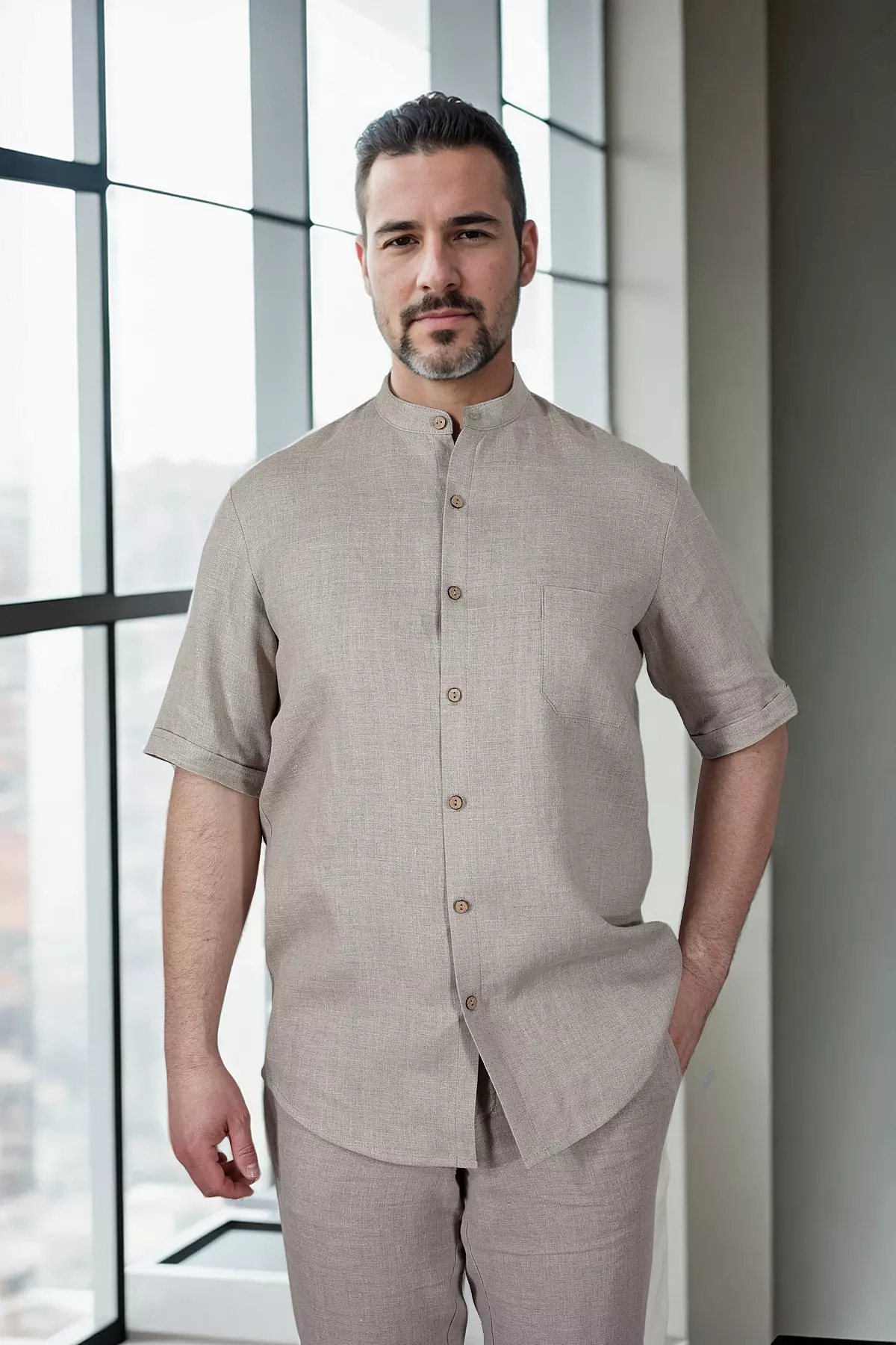 Mężczyzna stoi przodem na jasnym tle. Ma na sobie koszulę Tom w kolorze naturalnego lnu (piaskowym). Koszula ma stójkę, krótki rękaw i kieszonkę na piersi. Ręce trzyma w kieszeniach pasujących, lnianych spodni. Stylizacja jest spójna i letnia.