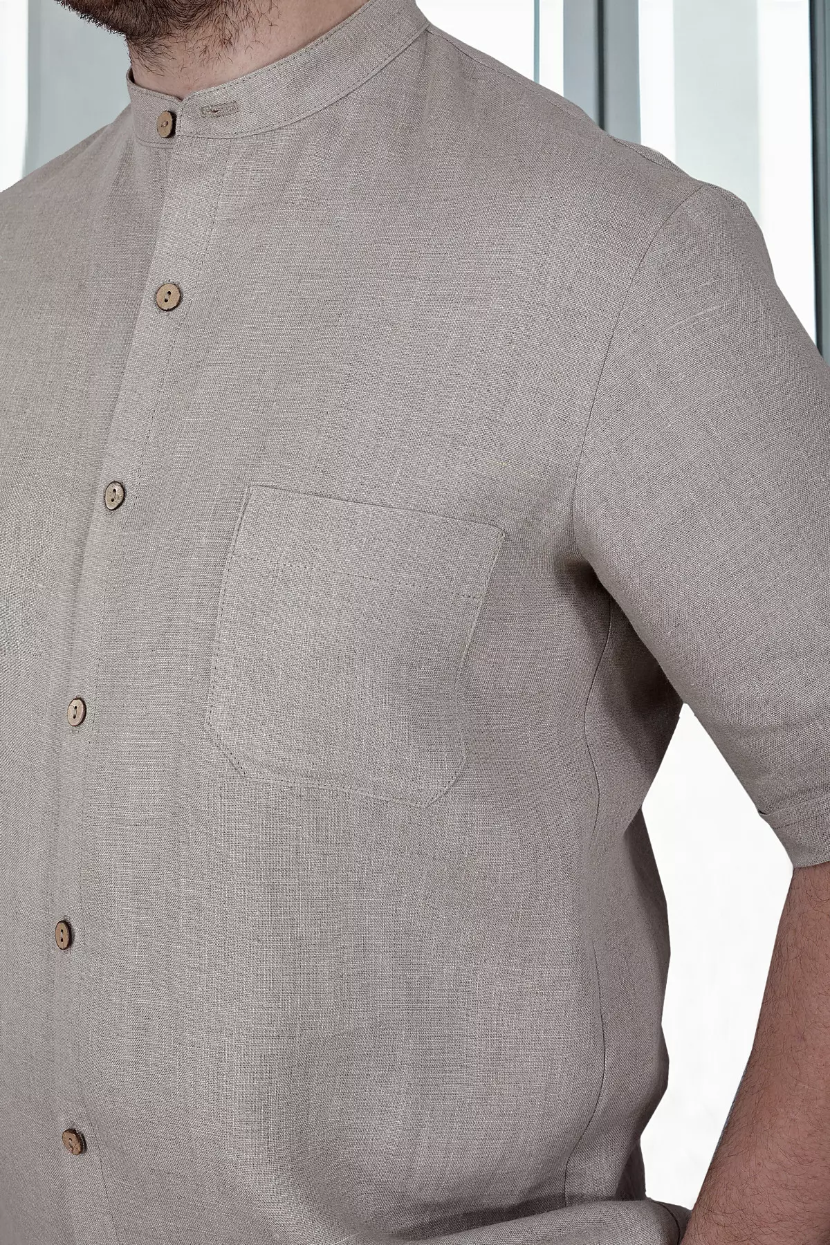 Zbliżenie na tors i szyję. Widać precyzyjnie wykończoną stójkę oraz kieszonkę na lewej piersi. Naturalna, surowa faktura lnu jest wyraźnie widoczna. Kontrastowe, brązowe guziki (drewniane lub kokosowe) dodają koszuli charakteru.