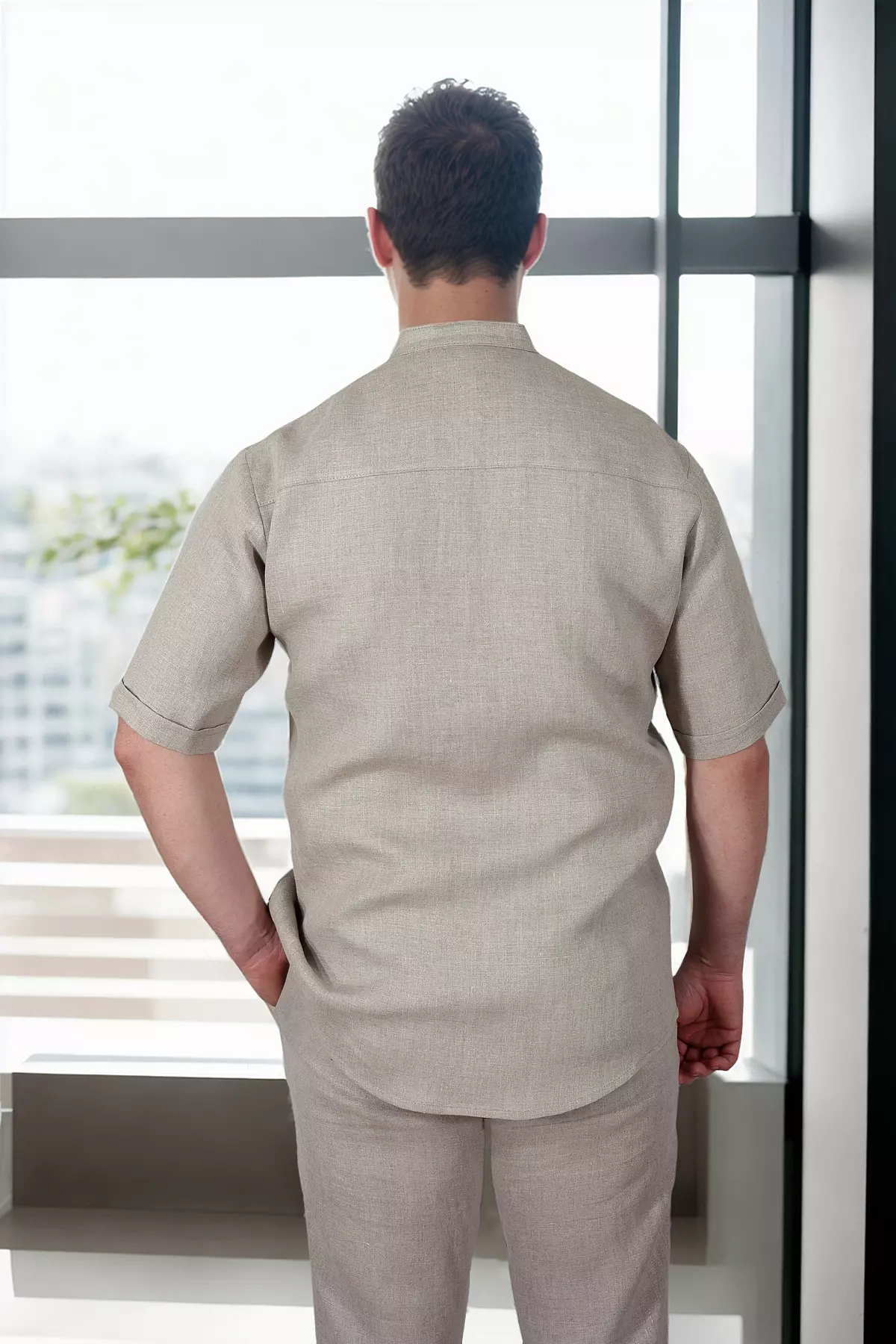Widok mężczyzny od tyłu, stojącego przy oknie. Koszula Tom ma prosty tył z poziomym karczkiem na łopatkach. Krótkie rękawy zapewniają swobodę. Kolor tkaniny to naturalny beż.