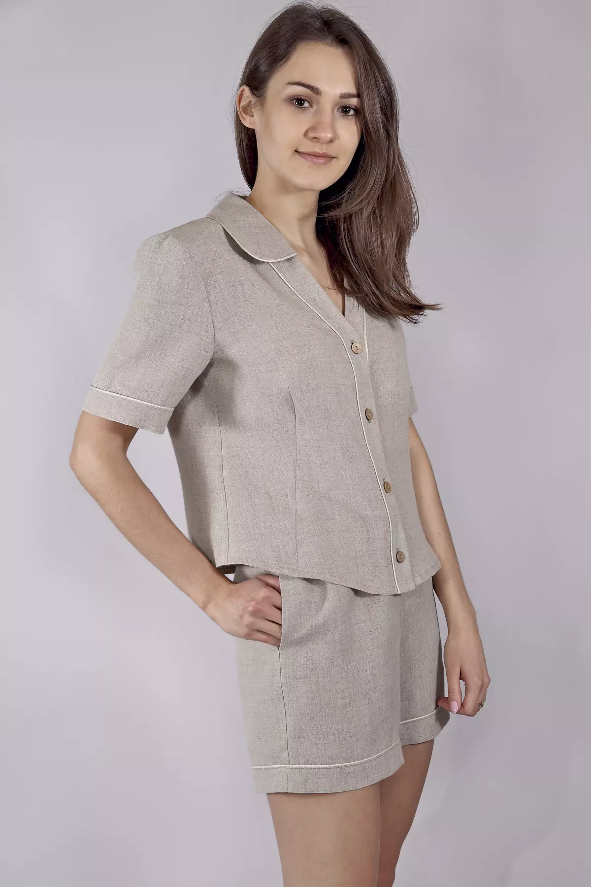 Modelka stoi w półprofilu, trzymając dłoń w kieszeni szortów. Naturalny, piaskowy odcień lnu harmonizuje z jasną wypustką przy krawędziach ubrania. Zestaw wygląda bardzo przytulnie i komfortowo.