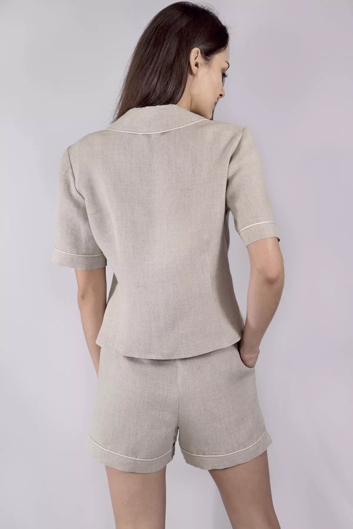 Widok pleców modelki. Prezentuje tył zestawu Kinia w kolorze naturalnego lnu. Widać koszulę z krótkim rękawem oraz szorty. Karczek, rękawy i nogawki wykończone są jasną, kontrastową wypustką. Krój jest luźny i swobodny.