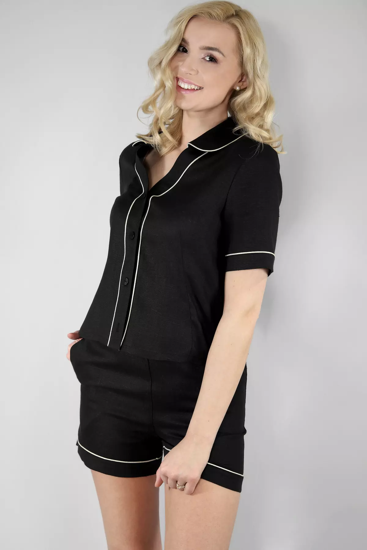 Uśmiechnięta modelka stoi w półprofilu. Ma na sobie czarną koszulę piżamową z kołnierzykiem i krótkie szorty. Jasna lamówka biegnie wzdłuż zapięcia, kołnierza i brzegów, tworząc elegancki kontur.