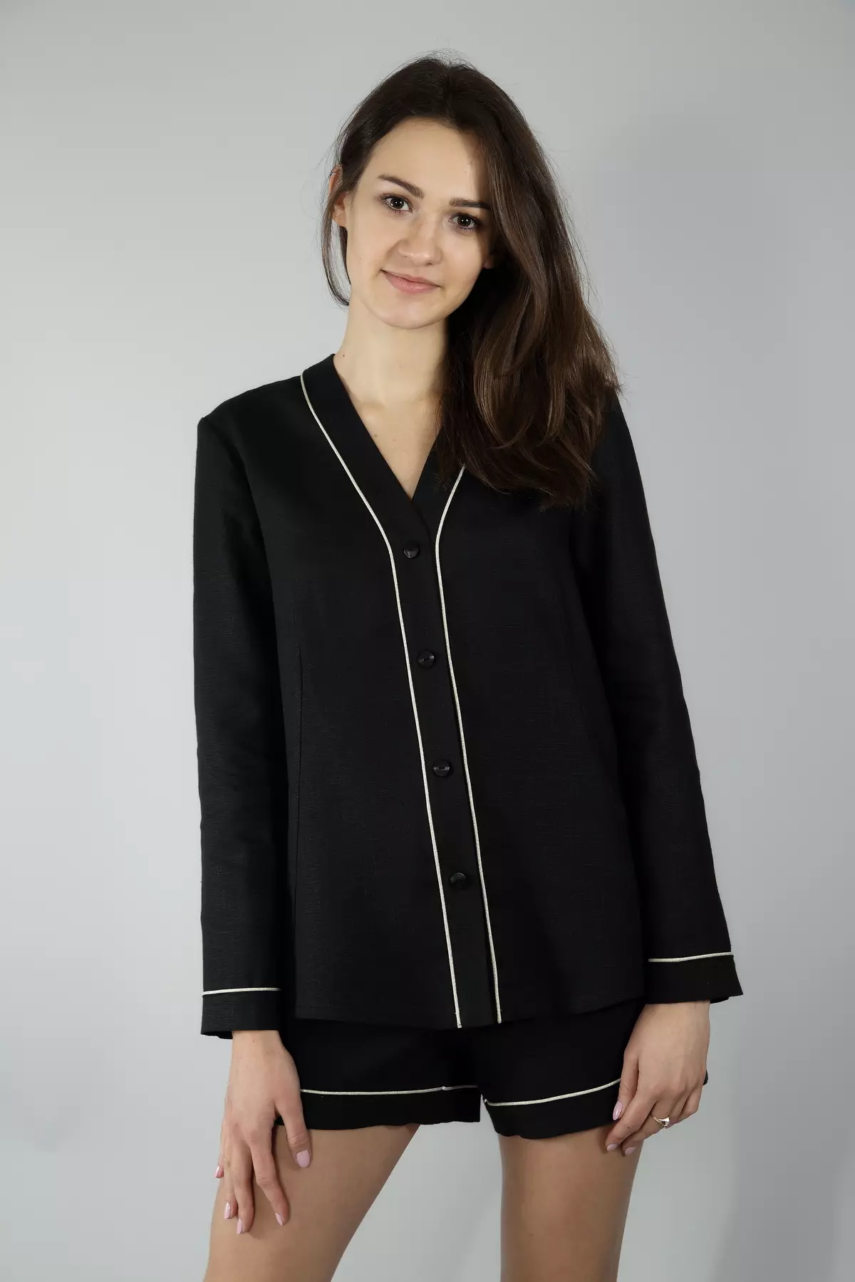 Modelka stoi prosto na szarym tle. Ubrana w czarną koszulę Daga zapinaną na czarne guziki oraz krótkie szorty. Dekolt V wysmukla szyję. Zestaw wygląda elegancko i nowocześnie.