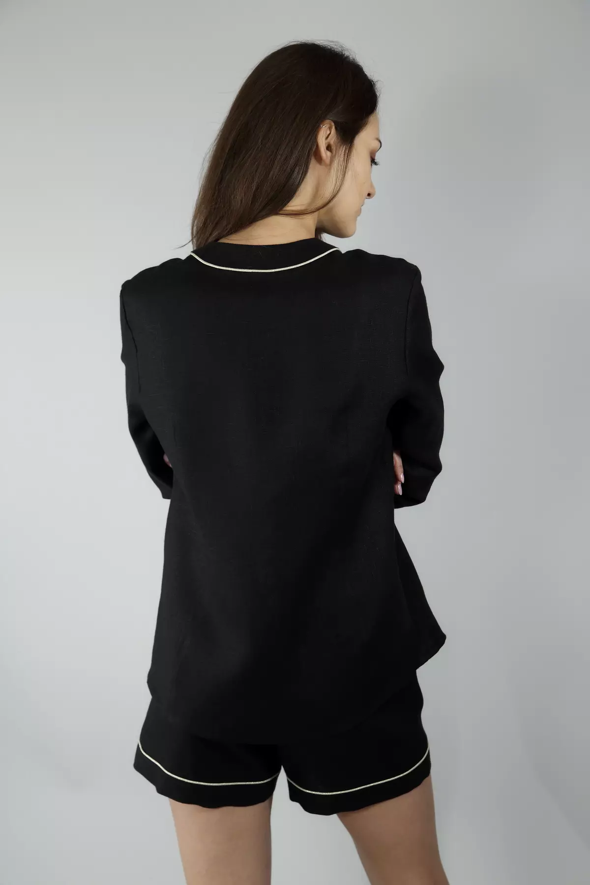 Widok pleców modelki. Czarna koszula Daga ma prosty tył. Widać, jak jasna wypustka biegnie wzdłuż dekoltu i dołu koszuli oraz przy mankietach i nogawkach szortów. Krój jest swobodny.