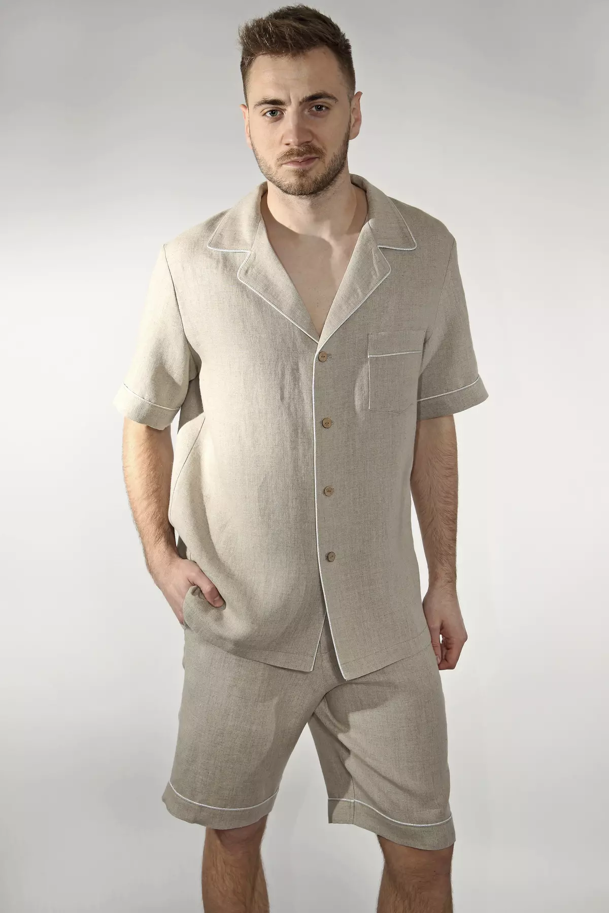Mężczyzna stoi przodem na jasnym tle. Ma na sobie piżamę Remi w kolorze naturalnego lnu (beżowym). Koszula z krótkim rękawem i kołnierzykiem zestawiona jest z krótkimi szortami. Jasna wypustka zdobi brzegi ubrania. Ręka mężczyzny w kieszeni szortów.