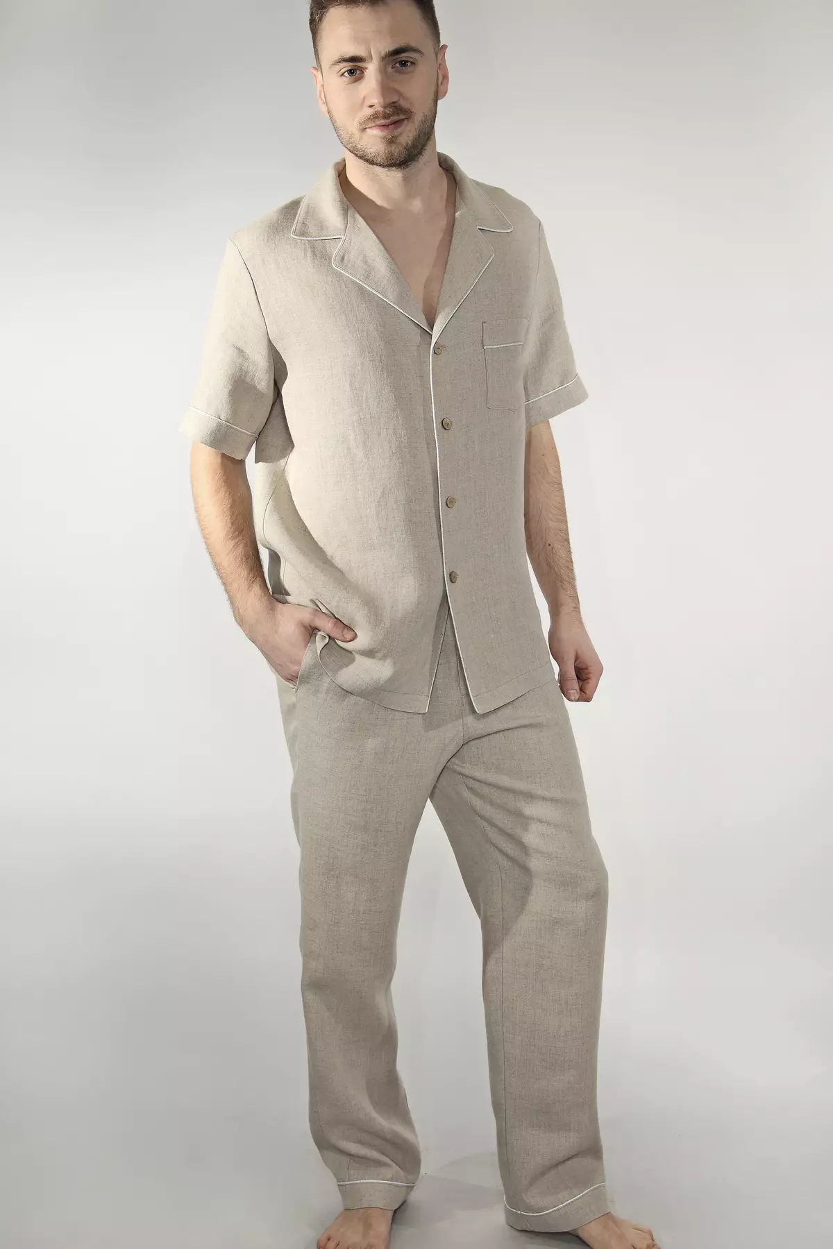 Mężczyzna stoi boso, w zestawie z długimi spodniami. Piaskowy odcień lnu i jasna wypustka tworzą harmonijną całość. Koszula z krótkim rękawem połączona z długimi spodniami to świetna opcja na okres przejściowy.