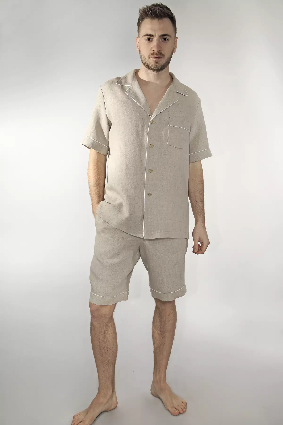 Mężczyzna stoi prosto, przodem do obiektywu. Prezentuje letni wariant piżamy Remi w kolorze naturalnego lnu: koszulę z krótkim rękawem i kołnierzykiem oraz krótkie spodenki. Prawa ręka modela jest schowana w kieszeni szortów, lewa swobodnie opuszczona. Wyraźnie widać jasną, kontrastową wypustkę wykańczającą krawędzie ubrania oraz naturalny, beżowy odcień tkaniny.