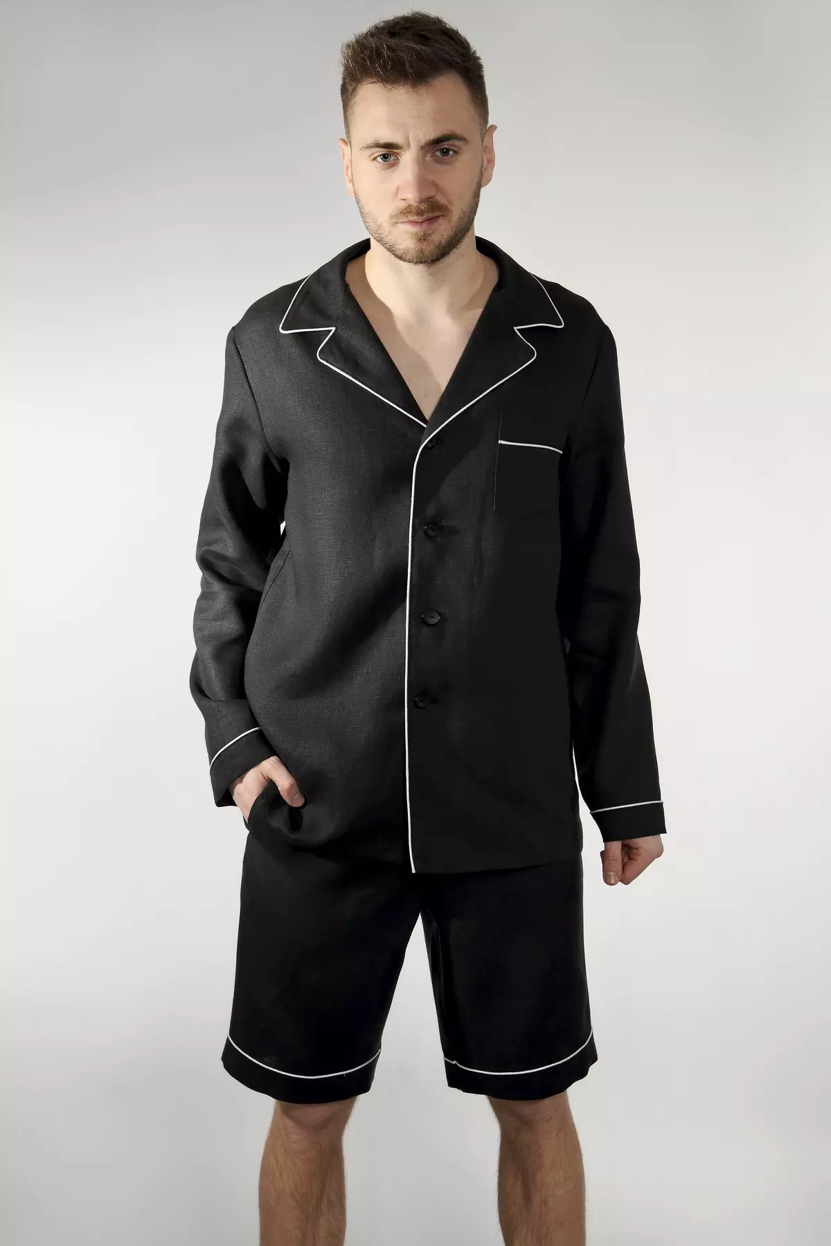 Mężczyzna stoi przodem w zestawie z szortami. Koszula z długim rękawem i kołnierzykiem nadaje stylizacji elegancji, a krótkie spodenki zapewniają przewiewność. Kontrastowa wypustka jest wyraźnym elementem ozdobnym.