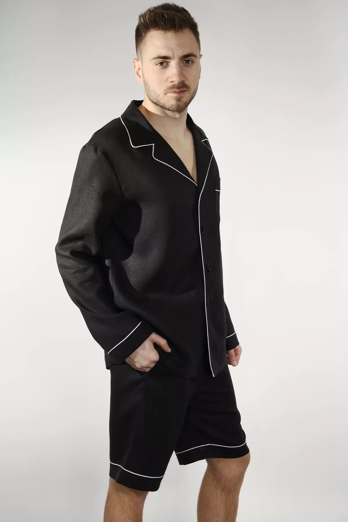 Mężczyzna stoi w półprofilu na jasnym tle. Prezentuje czarną piżamę Axel składającą się z koszuli z długim rękawem i szortów. Koszula ma kołnierzyk i jest wykończona jasną, kontrastową wypustką. Mężczyzna trzyma rękę w kieszeni szortów.