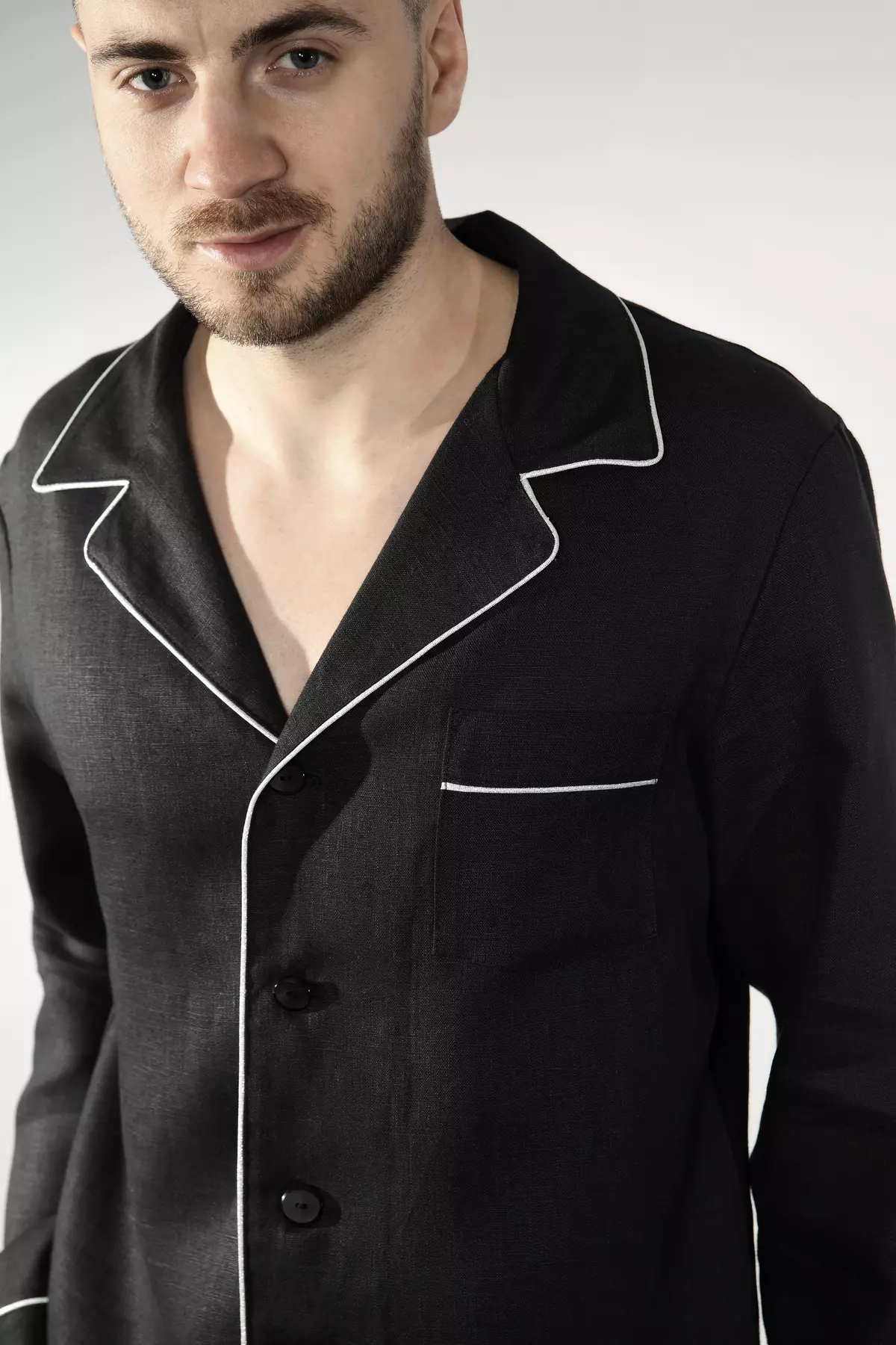 Zbliżenie na tors i twarz modela. Mężczyzna uśmiecha się delikatnie. Widać precyzyjne wykończenie kołnierzyka i kieszonki na piersi białą wypustką. Czarny len ma matową, szlachetną fakturę.