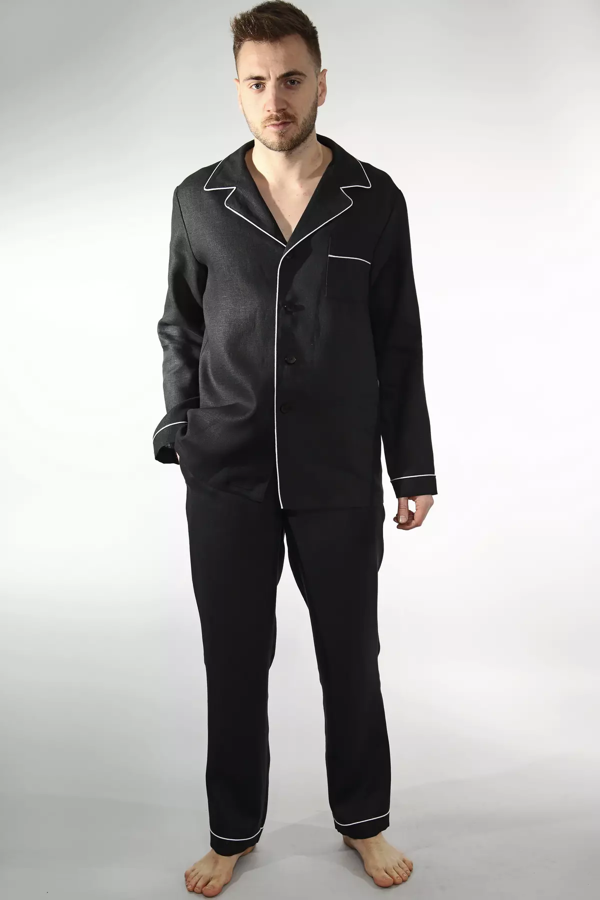 Mężczyzna stoi boso, w pełnym zestawie z długimi spodniami. Czarna, lniana piżama wygląda bardzo elegancko. Jasna wypustka przy kołnierzu, mankietach i nogawkach dodaje jej charakteru. Koszula jest zapięta na guziki.