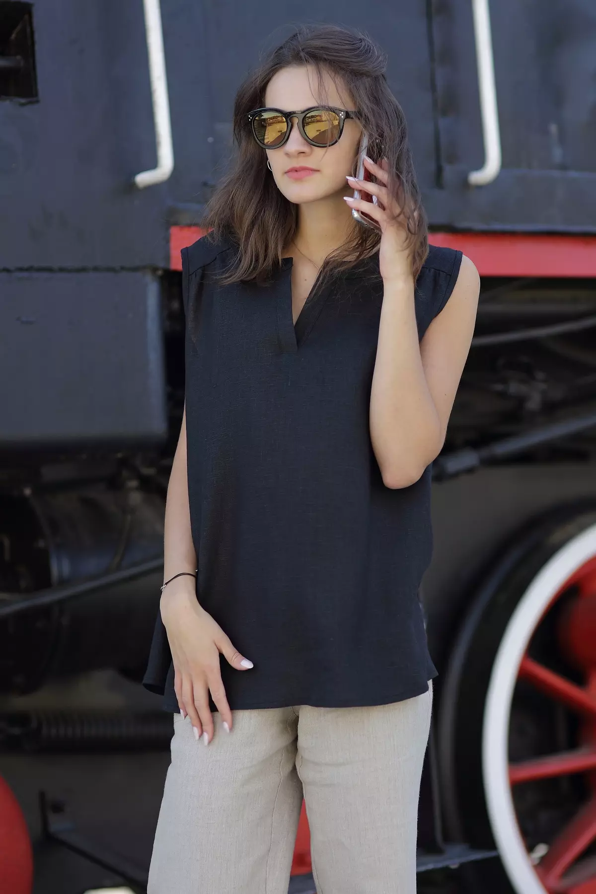 Modelka rozmawia przez telefon. Ma na sobie czarną bluzkę Rena, która świetnie sprawdza się w stylizacji do pracy. Widać eleganckie wykończenie dekoltu w serek z pionową plisą. Czerń bluzki kontrastuje z beżem spodni.
