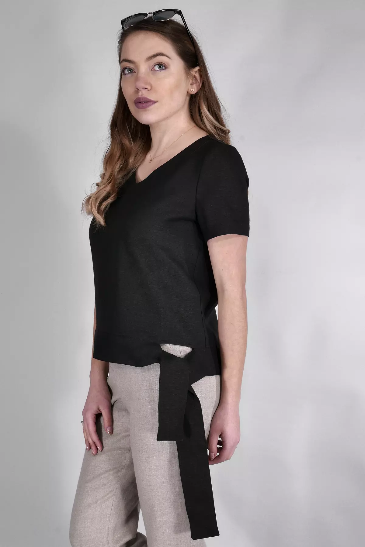 Modelka stoi w półprofilu na jasnym tle. Ma na sobie czarną bluzkę Nika z krótkim rękawem. Wyróżnia się ona szeroką, czarną szarfą wiązaną na boku biodra. Modelka ma okulary przeciwsłoneczne założone na włosy.