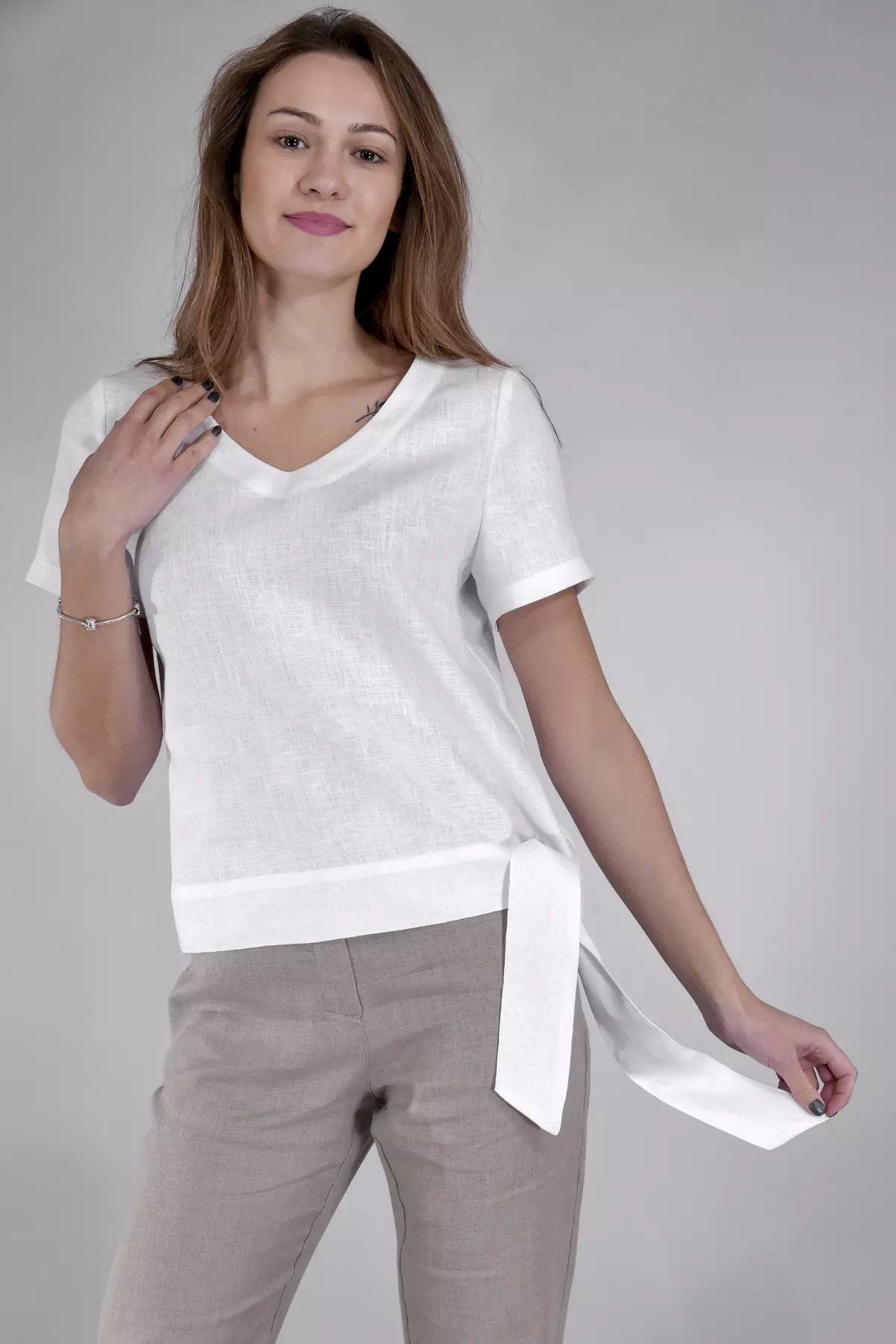 Modelka stoi w półprofilu, dotykając dłonią dekoltu. Ma na sobie białą bluzkę Nika. Charakterystyczne wiązanie na boku (szeroka szarfa) jest wyraźnie widoczne. Bluzka zestawiona jest z beżowymi spodniami.