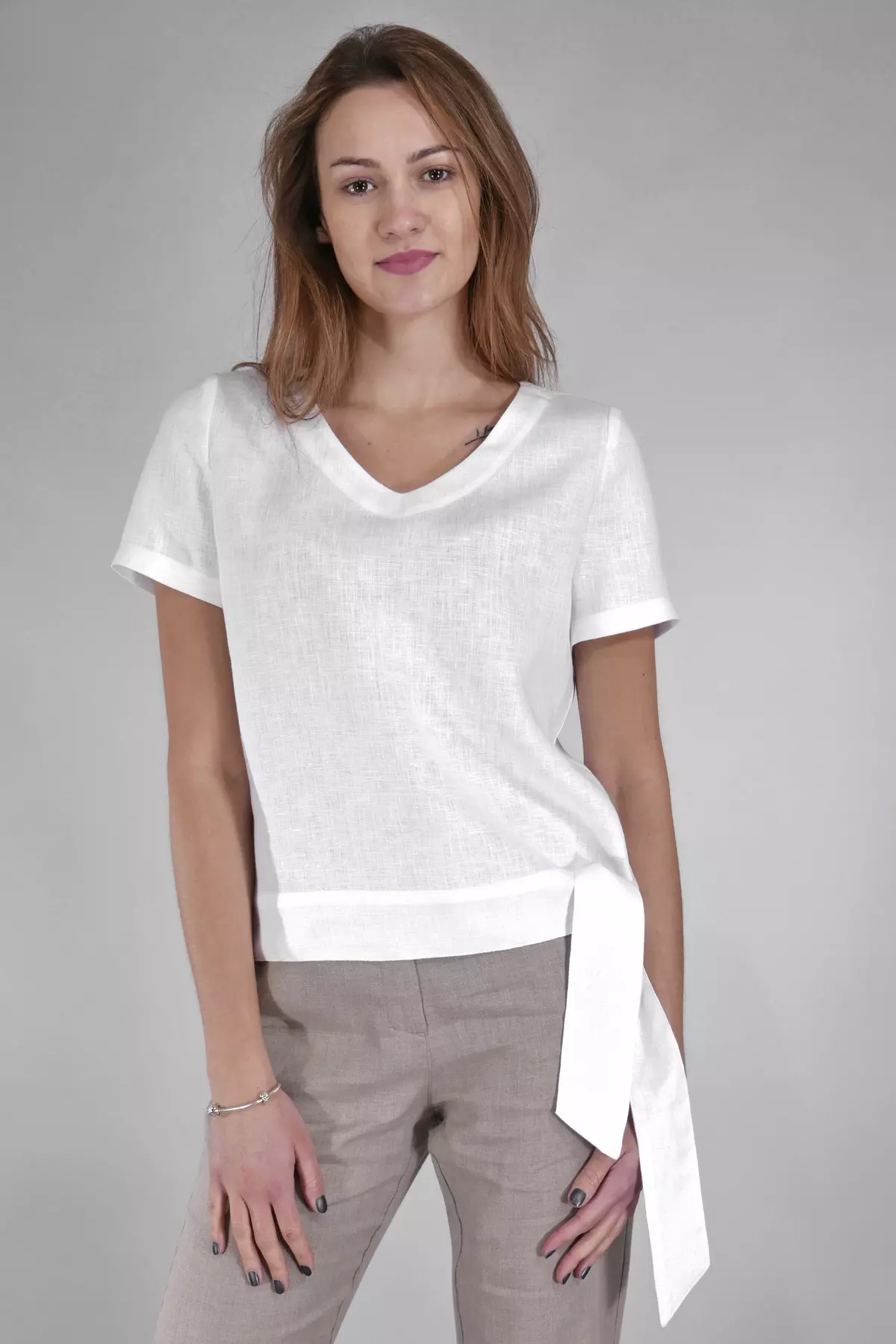 Modelka stoi prosto na szarym tle. Prezentuje białą bluzkę Nika z wiązaniem na boku. Dekolt w serek i krótki rękaw nadają jej lekkości. Wiązanie tworzy asymetryczny dół, który dodaje stylizacji dynamiki.