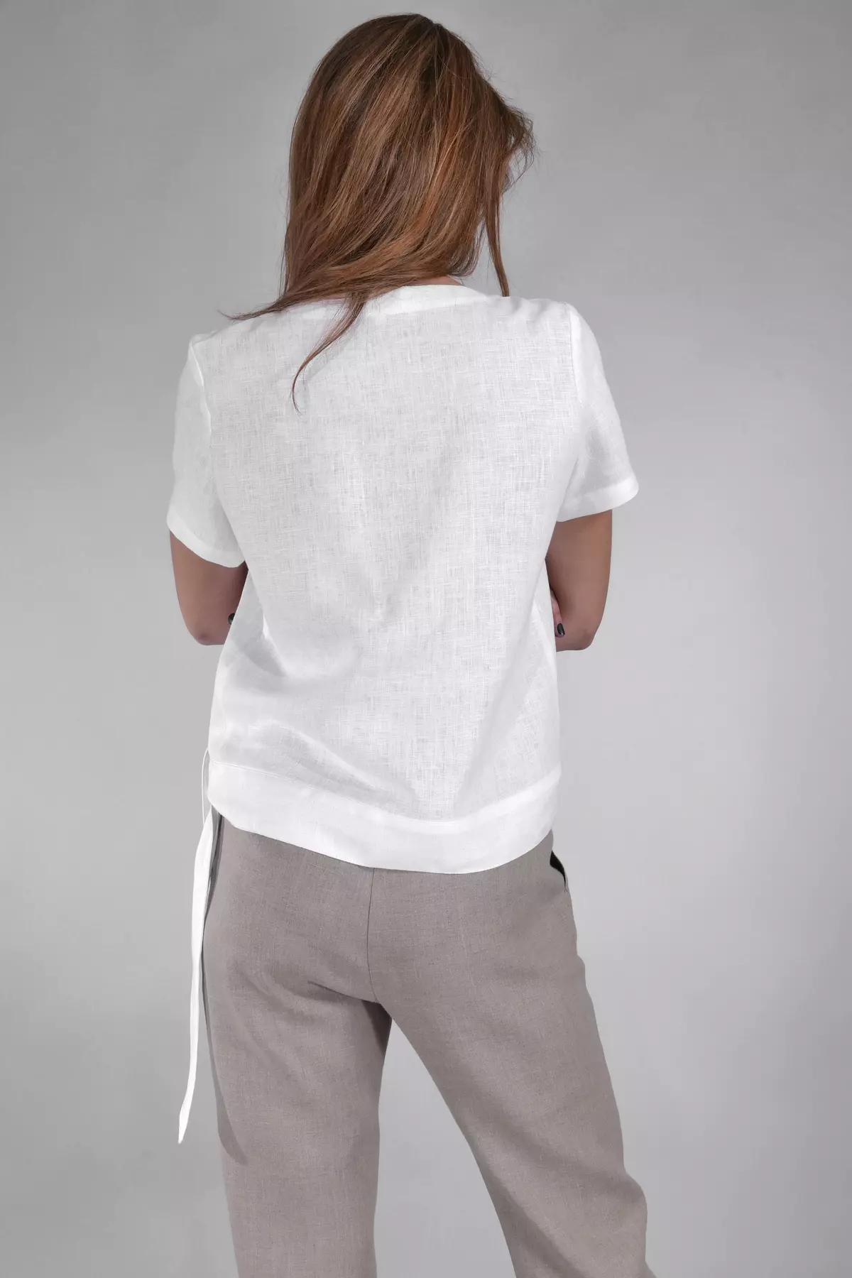 Widok pleców modelki. Biała bluzka Nika ma prosty, gładki tył. Po lewej stronie widać długą, białą szarfę zwisającą luźno wzdłuż biodra. Krój bluzki jest swobodny, nie opina sylwetki.