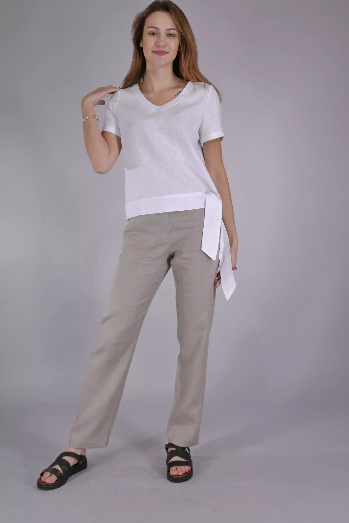 Uśmiechnięta modelka stoi w swobodnej pozie, poprawiając włosy. Ma na sobie białą bluzkę Nika i beżowe, lniane spodnie. Szarfa wiązana na boku jest rozwiązana i luźno opada, co wygląda bardzo efektownie.
