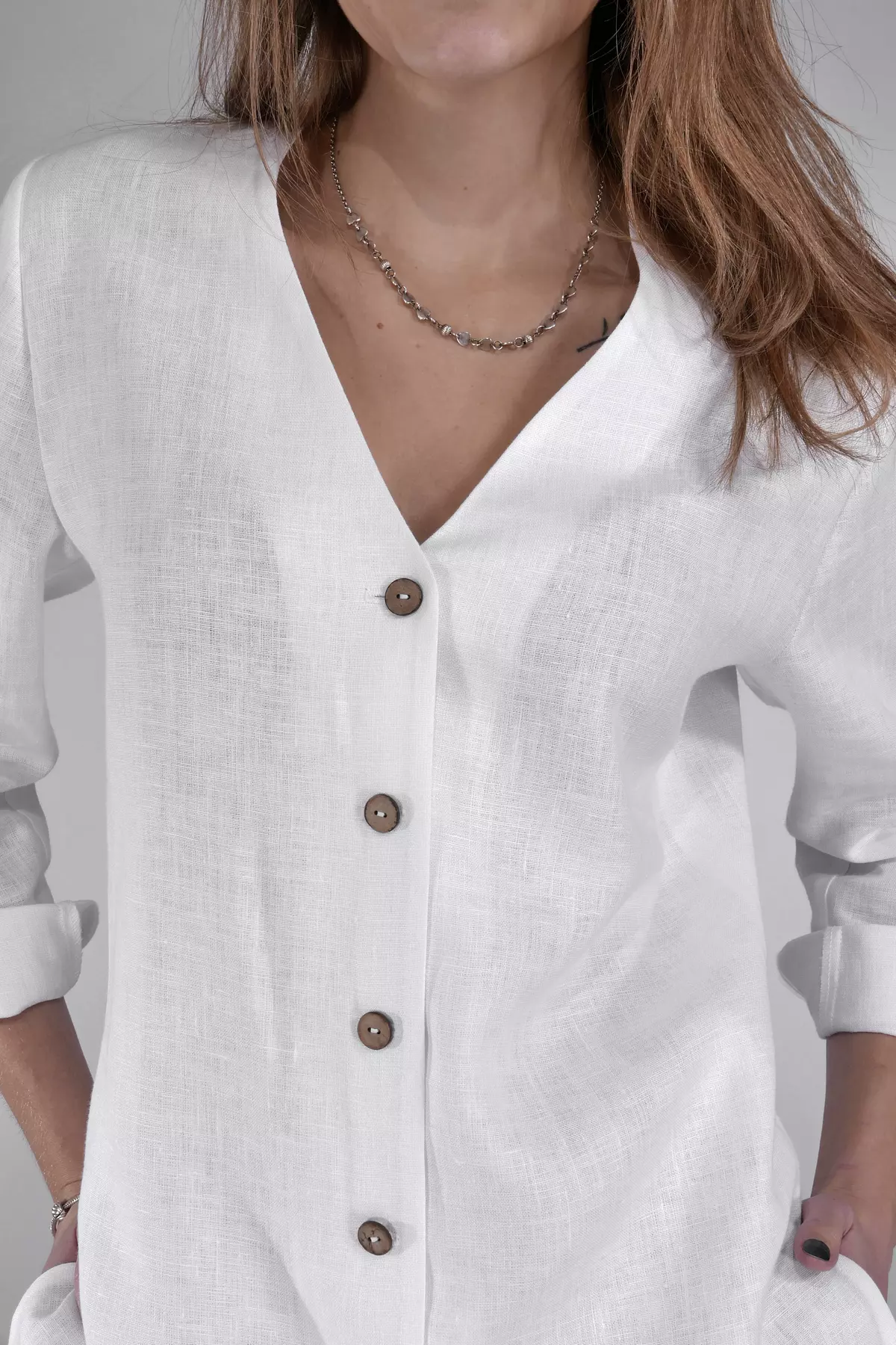 Zbliżenie na górną część bluzki. Widać wykończenie dekoltu w kształcie litery V (brak kołnierzyka) oraz fakturę lnianej tkaniny. Naturalne, ciemniejsze guziki stanowią ozdobny akcent na tle bieli.