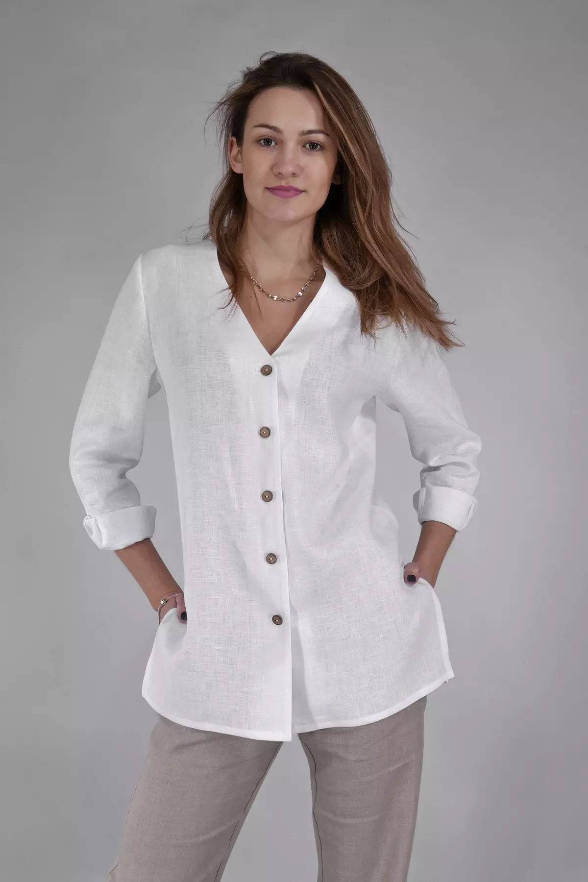 Modelka stoi przodem, trzymając ręce w kieszeniach beżowych spodni. Prezentuje białą bluzkę Lara. Wyróżnia się ona dekoltem w serek bez kołnierzyka oraz rzędem kontrastowych, brązowych guzików.