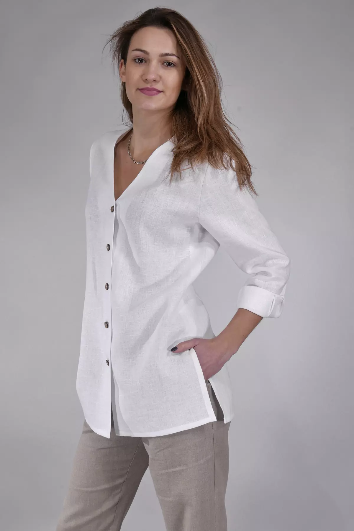 Modelka stoi bokiem, wkładając rękę do kieszeni spodni. Bluzka Lara swobodnie opada na biodra. Długie rękawy są proste i eleganckie. Stylizacja łączy biel koszuli z naturalnym beżem spodni.