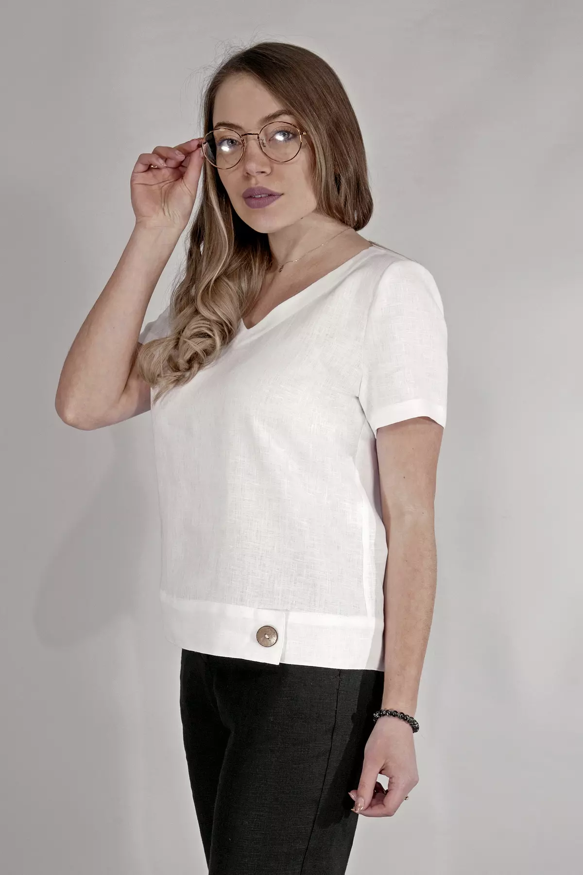 Modelka w okularach korekcyjnych poprawia włosy. Ubrana w białą, lnianą bluzkę Carla i czarne spodnie. Stylizacja jest biurowa, ale swobodna. Widać fakturę białego lnu i precyzyjne wykończenie dekoltu.