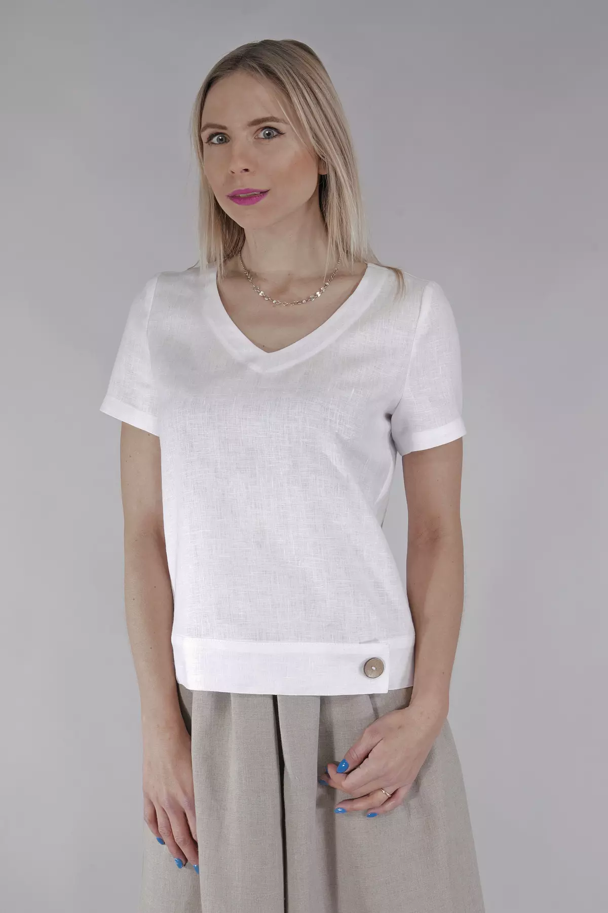 Blondynka stoi prosto na szarym tle. Ma na sobie białą bluzkę Carla o luźnym, pudełkowym kroju. Bluzka ma krótki rękaw i dekolt w kształcie litery V. Na dole, po lewej stronie, widać charakterystyczny drewniany guzik. Zestawiona jest z beżową, lnianą spódnicą.
