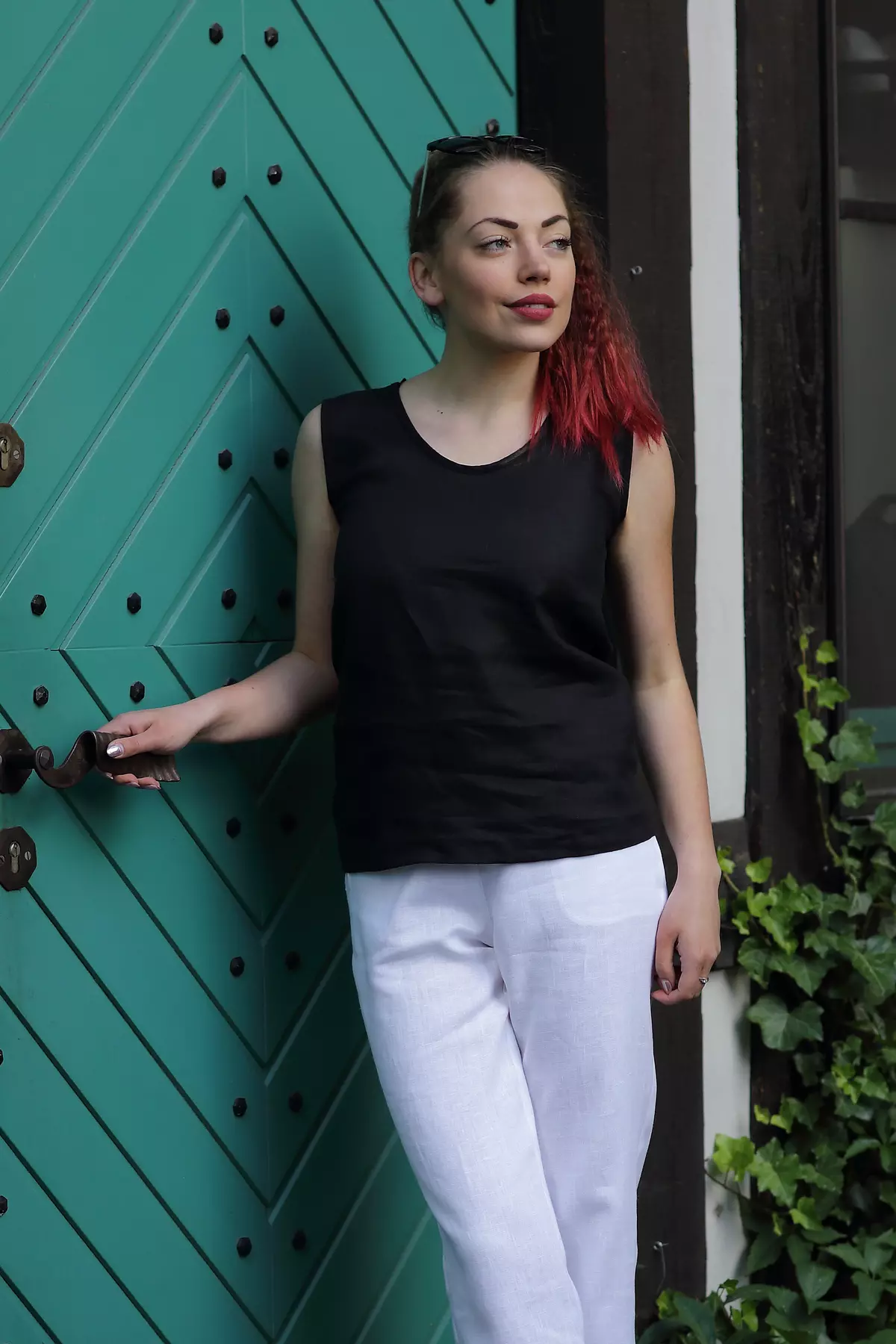 Modelka stoi przy dużych, zielonych, drewnianych drzwiach. Jedną ręką trzyma za klamkę. Ma na sobie czarną bluzkę Bea, która luźno opada na białe spodnie. Czerń bluzki jest głęboka i matowa.