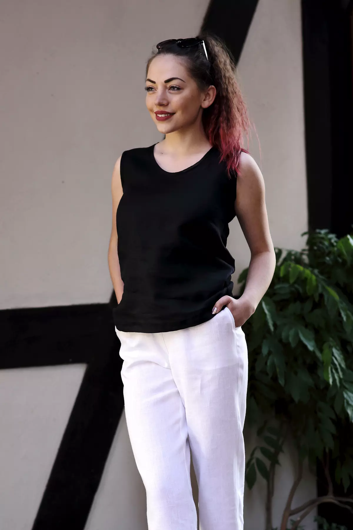 Uśmiechnięta brunetka z czerwonymi końcówkami włosów stoi na tle budynku z muru pruskiego. Nosi czarny top Bea i białe, lniane spodnie. Ręce trzyma w kieszeniach. Stylizacja jest kontrastowa i letnia.