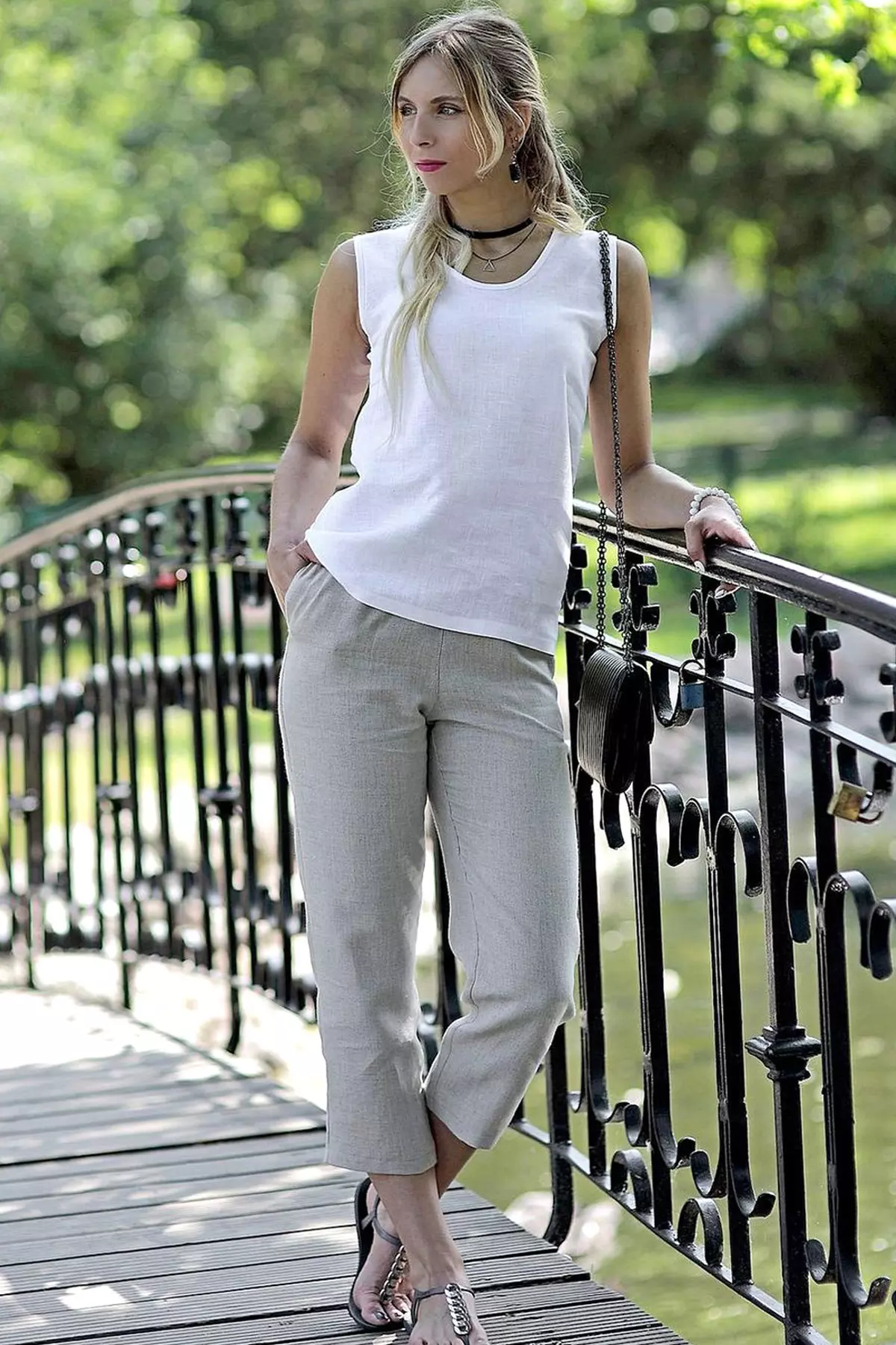 Blondynka stoi na mostku w parku, opierając się o barierkę. Ma na sobie biały top Bea i beżowe rybaczki. Stylizacja uzupełniona jest o choker i małą torebkę na łańcuszku. Słońce oświetla biały materiał.