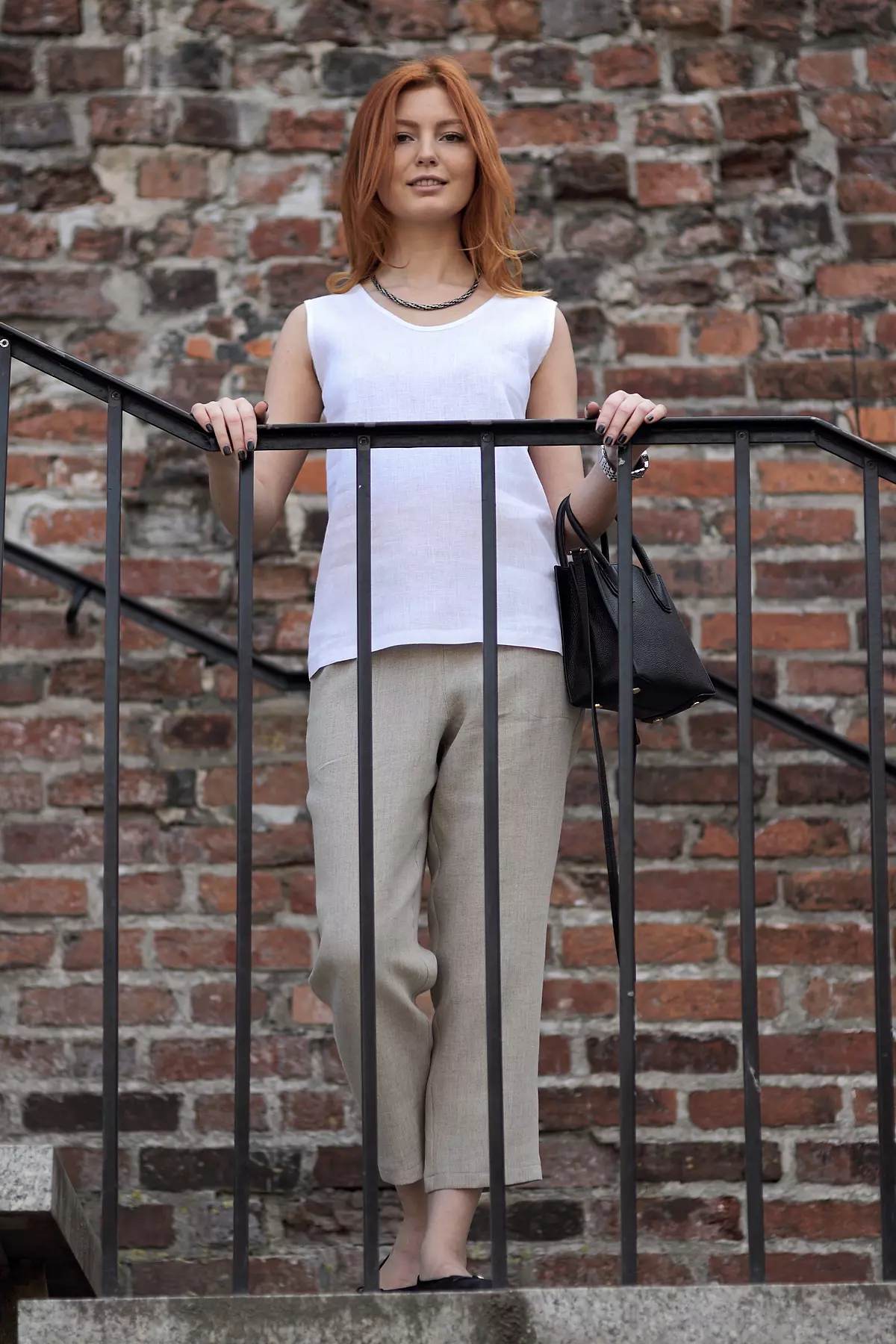Ruda modelka stoi na schodach przy metalowej barierce, na tle ceglanego muru. Ma na sobie białą bluzkę Bea bez rękawów o luźnym kroju. Zestawiła ją z beżowymi, lnianymi spodniami i czarną torebką.