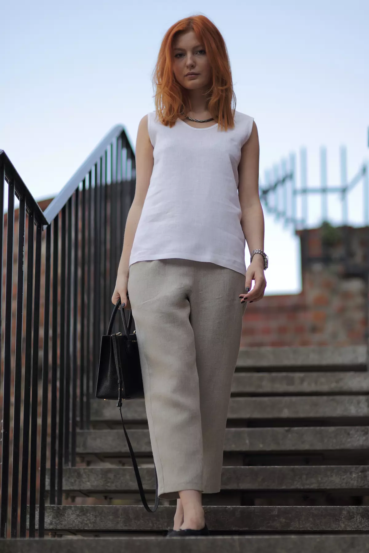 Modelka schodzi po betonowych schodach. Biała bluzka Bea swobodnie opływa sylwetkę. Prosty, okrągły dekolt i brak rękawów sprawiają, że jest to idealny wybór na lato. W ręku trzyma czarną torebkę.
