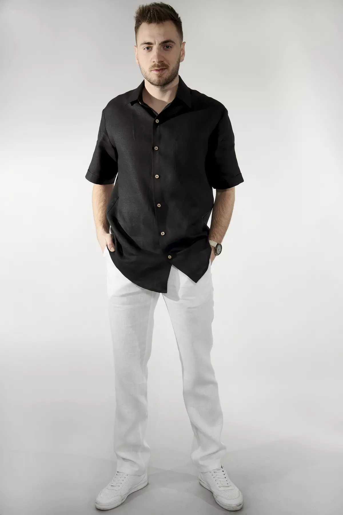 Mężczyzna uśmiecha się lekko, stojąc przodem. Ręce trzyma w kieszeniach. Czarna koszula Goran z krótkim rękawem i kołnierzykiem wygląda na wygodną i przewiewną. Kontrastowe guziki są wyraźnym elementem ozdobnym.