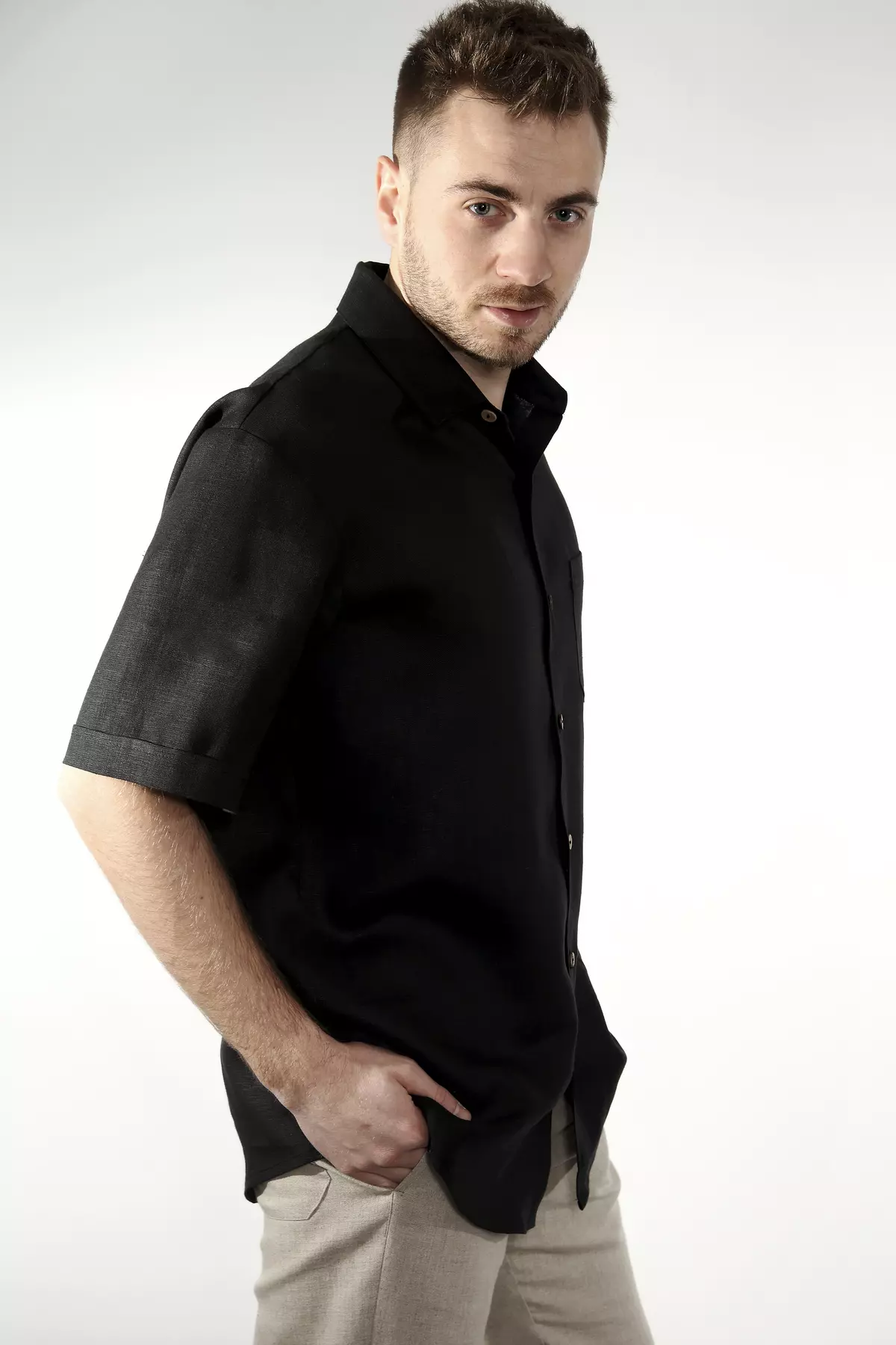 Mężczyzna stoi bokiem do obiektywu, patrząc w jego stronę. Ubrany w czarną koszulę Goran i beżowe spodnie. Ręka w kieszeni. Krój koszuli jest swobodny, ale elegancki. Czerń lnu ma matowe wykończenie.