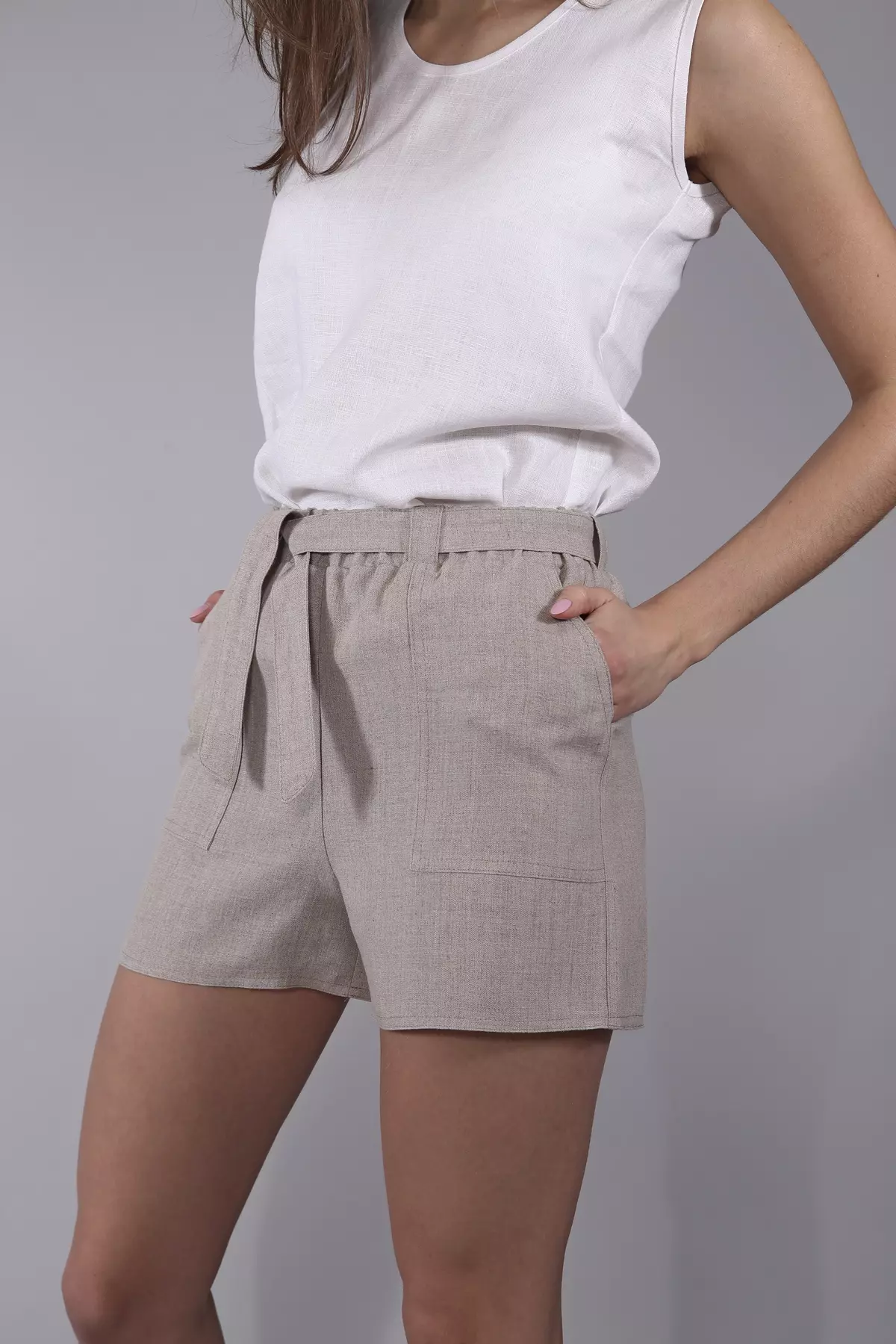 Modelka stoi w półprofilu, trzymając dłonie w kieszeniach. Luźna nogawka szortów zapewnia swobodę. Naturalny kolor spodenek harmonizuje z białą bluzką. Stylizacja jest lekka i ekologiczna.