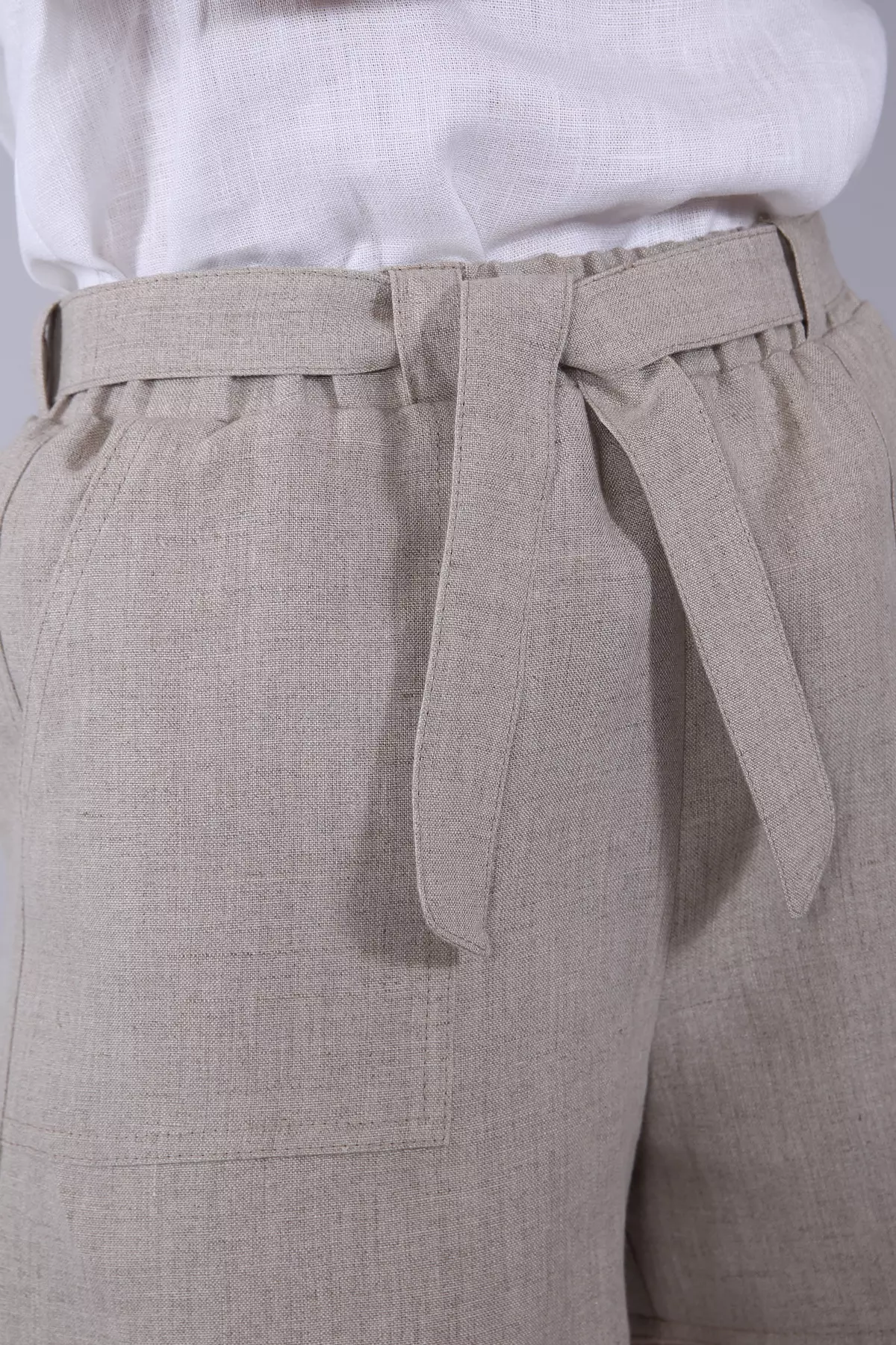 Zbliżenie na biodro i talię. Widać dłoń modelki w kieszeni. Zdjęcie ukazuje surową, piękną fakturę niebarwionego lnu oraz detale wykończenia: gumkę w pasie i szeroki pasek do wiązania.