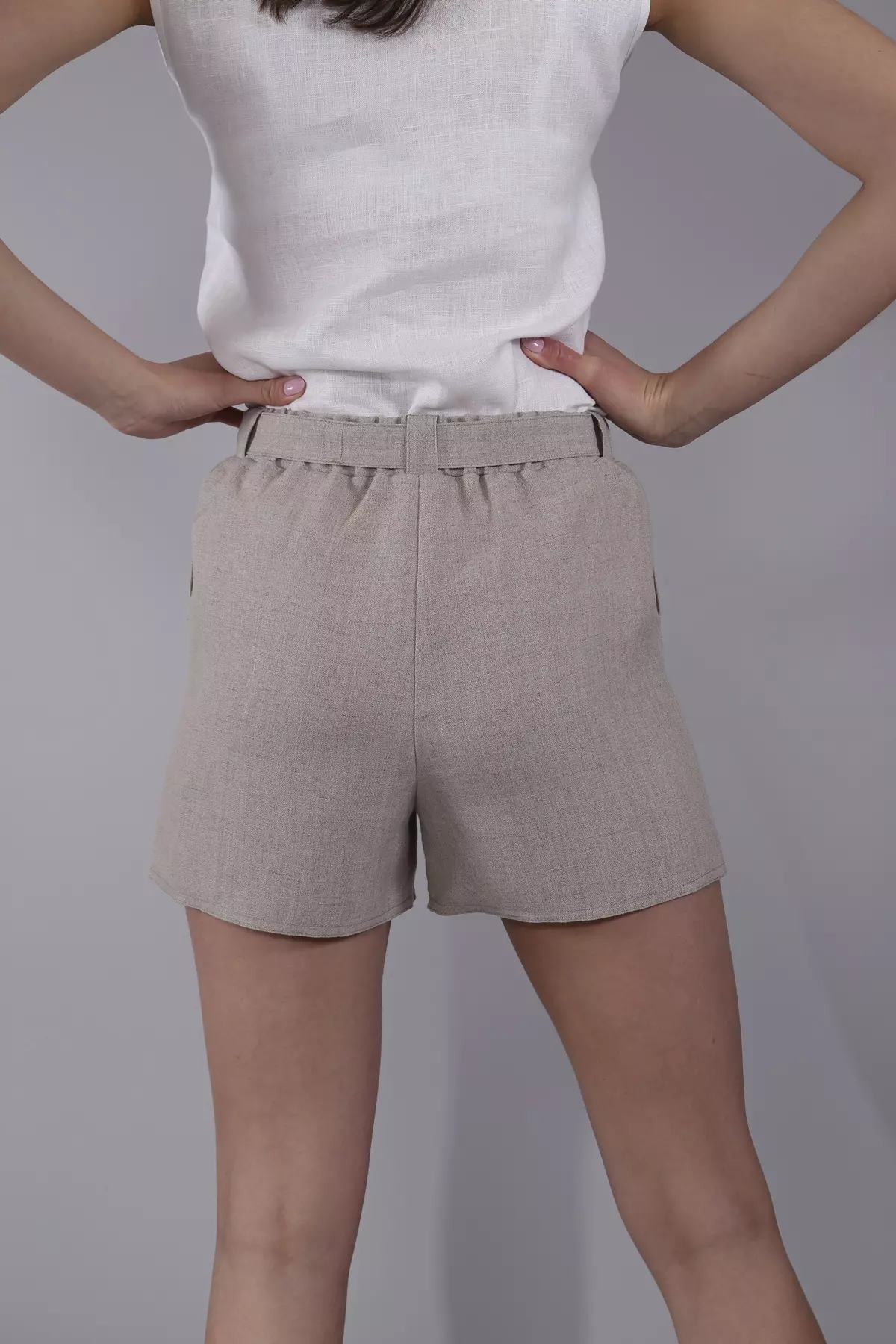 Widok szortów od tyłu. Modelka stoi w lekkim rozkroku, ręce oparte na biodrach. Szorty Maja w kolorze naturalnego lnu mają luźny krój, który nie opina sylwetki. Widać marszczenie przy elastycznej gumce w pasie oraz szlufki.