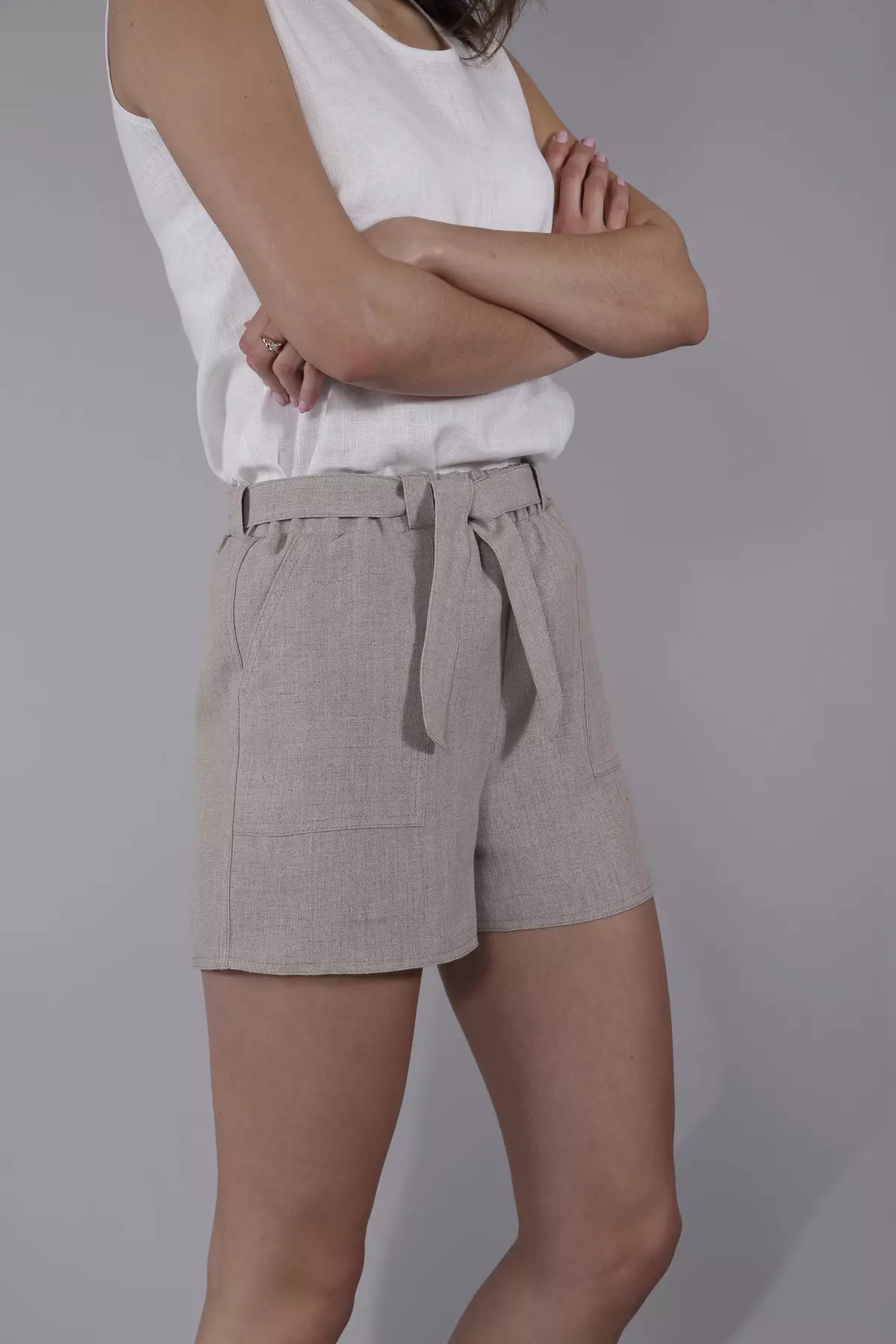 Modelka stoi bokiem ze skrzyżowanymi rękami. Ma na sobie beżowe szorty Maja i biały, lniany top. W pasie widoczne jest wiązanie na supeł z tego samego materiału. Spodenki mają duże, naszywane kieszenie.