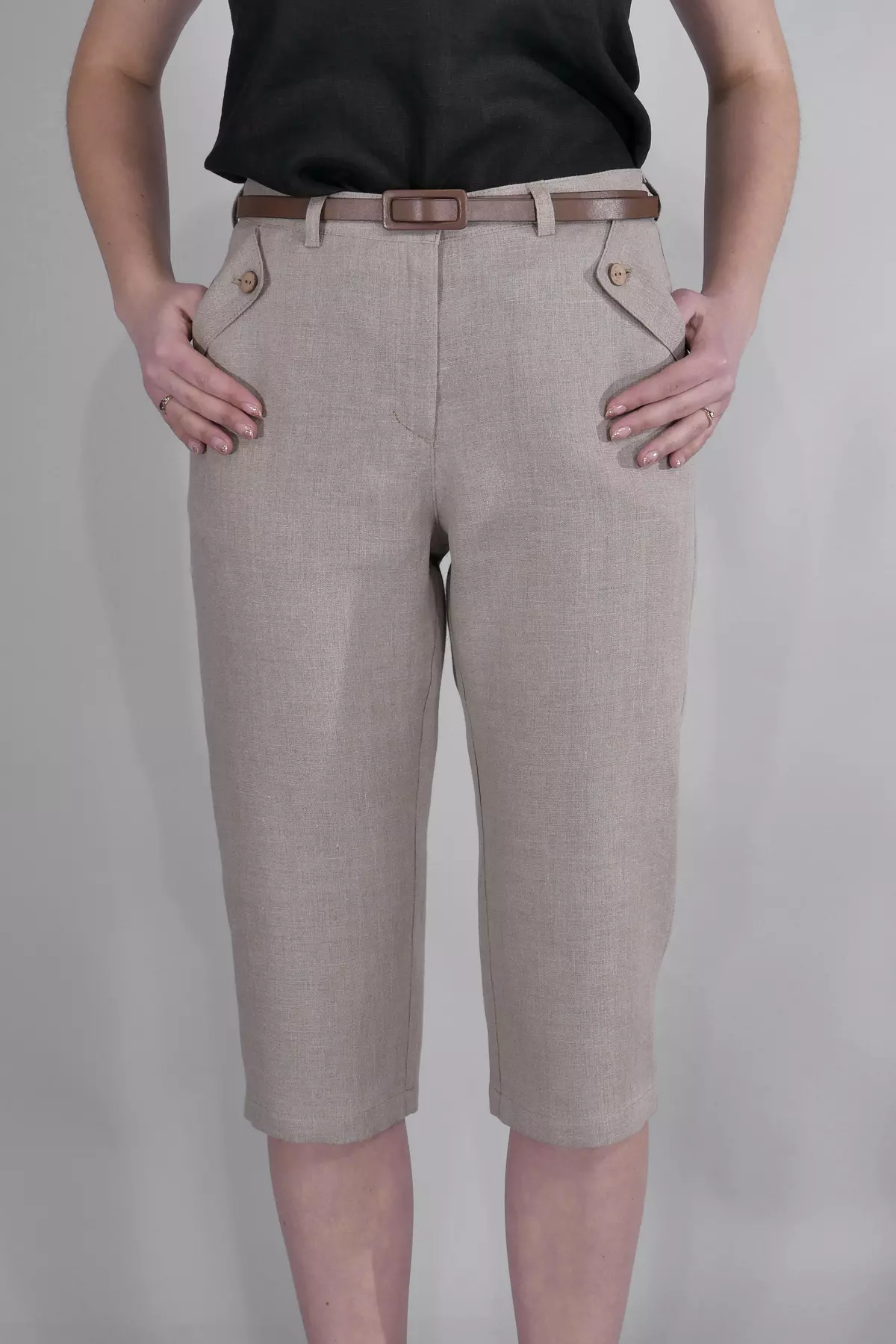 Modelka stoi przodem, trzymając kciuki w kieszeniach. Spodnie Nata w kolorze beżowym mają charakterystyczne, trójkątne patki przy kieszeniach zapinane na guziki. Widać szlufki i zapięcie na guzik w pasie.