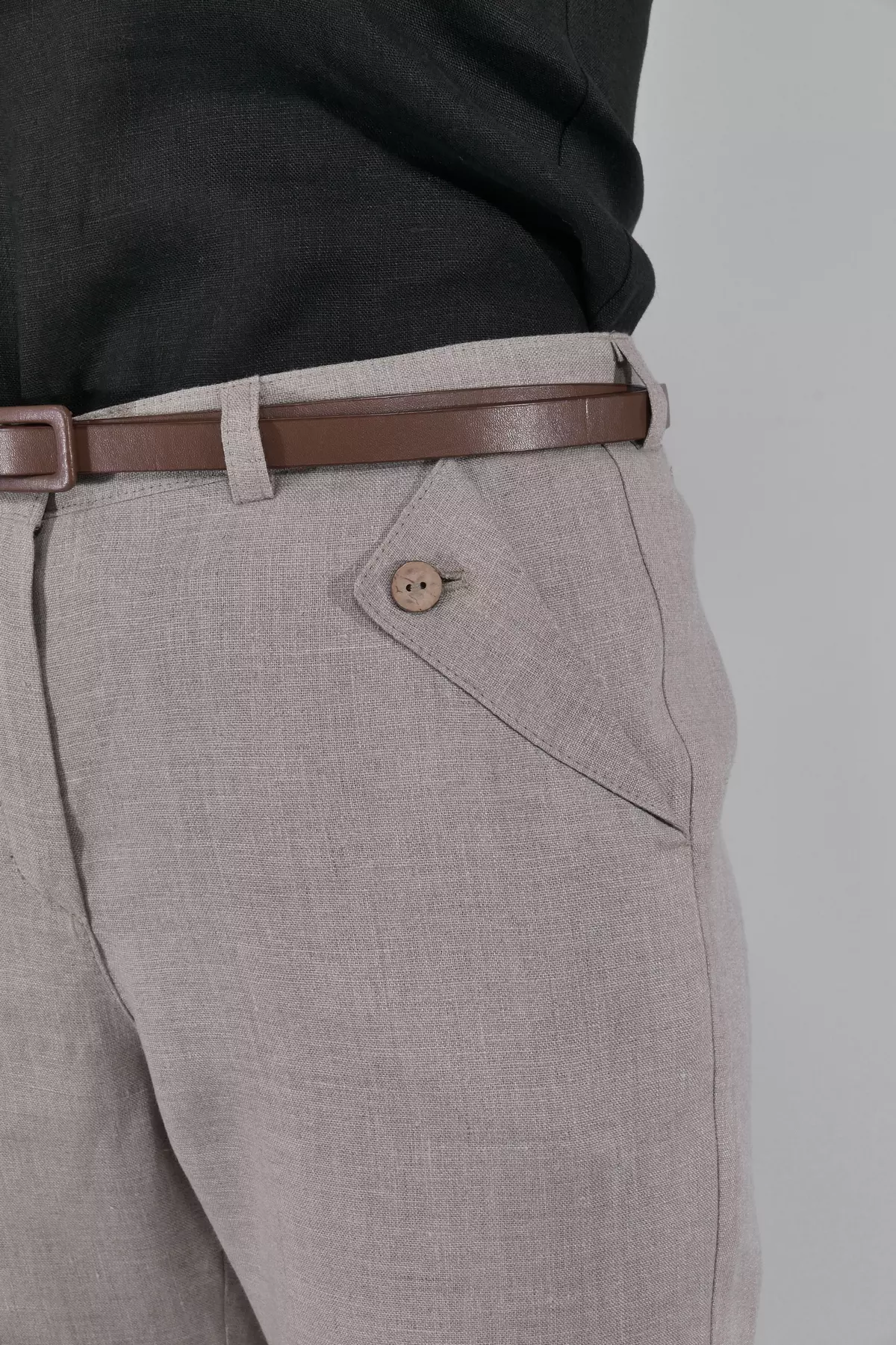 Zbliżenie na biodro i kieszeń. Widać surowy splot lnianej tkaniny w kolorze owsianym oraz brązowy, drewniany guzik przy kieszeni. Brązowy pasek idealnie komponuje się z całością.