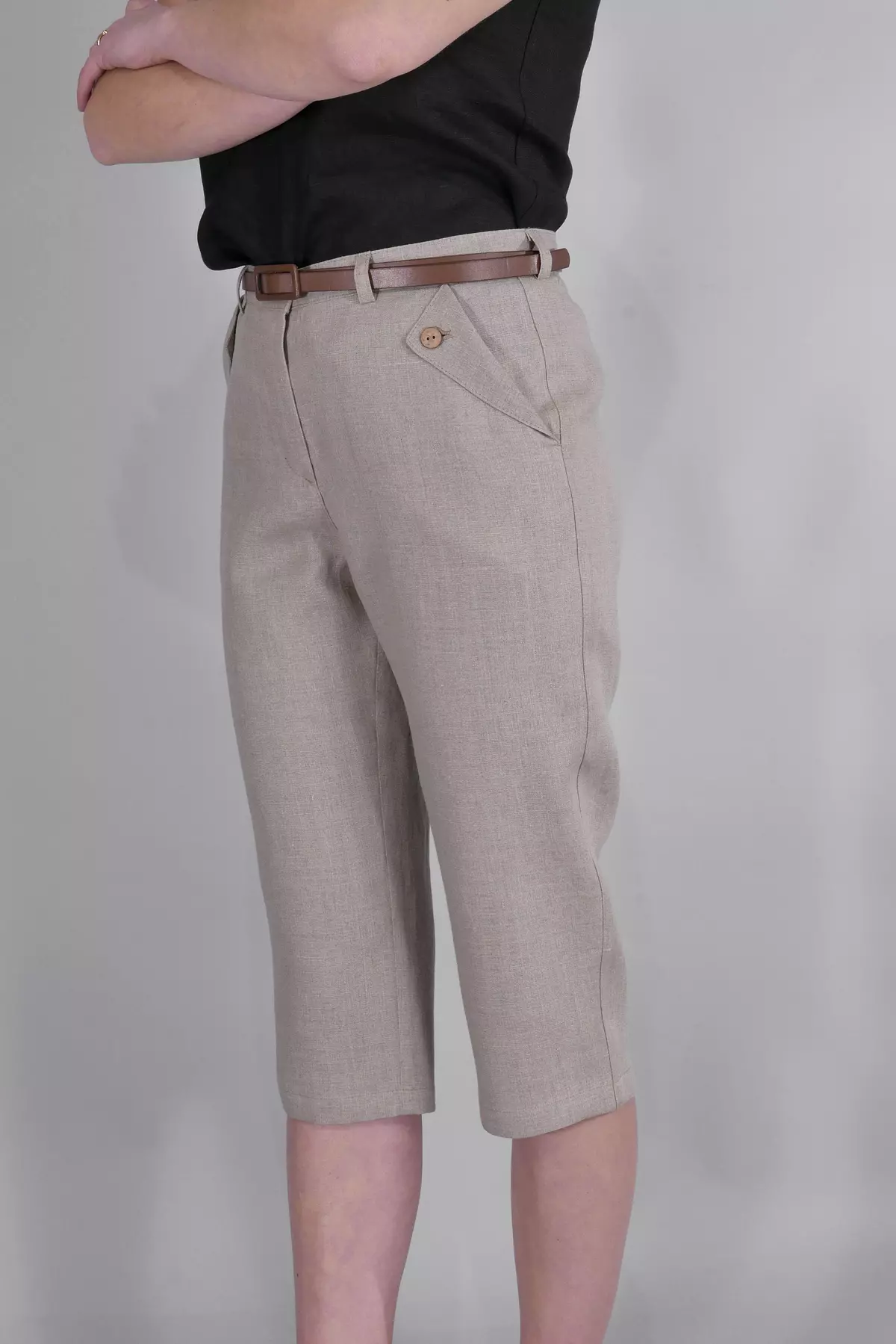 Modelka stoi w półprofilu, krzyżując ręce na klatce piersiowej. Prezentuje spodnie Nata w kolorze naturalnego lnu. Spodnie mają długość 3/4 i ozdobne kieszenie z guzikami. Stylizacja uzupełniona jest brązowym paskiem.