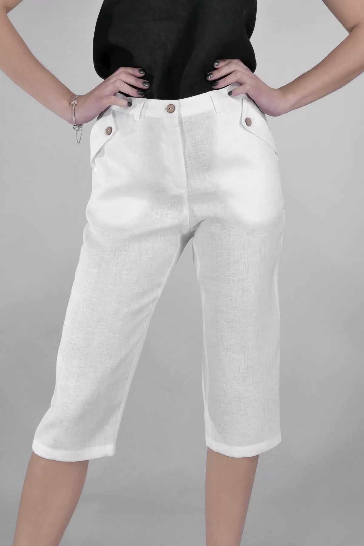 Modelka stoi pewnie z rękami na biodrach. Prezentuje białe rybaczki Nata. Widać zapięcie na guzik w pasie, szlufki oraz ozdobne patki przy kieszeniach. Spodnie są idealne na lato.