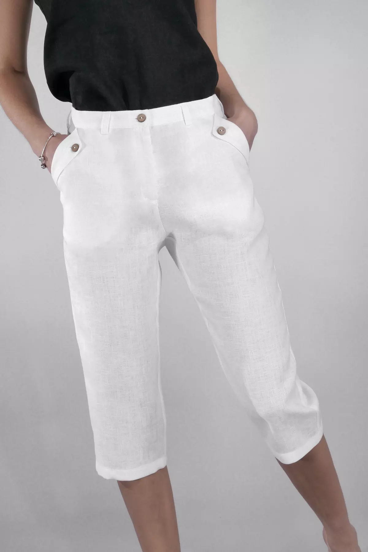 Zbliżenie na białe spodnie lniane Nata. Modelka trzyma dłonie w kieszeniach, które są ozdobione patkami zapinanymi na brązowe guziki. Spodnie mają szlufki w pasie i są zapinane na guzik. Góra stylizacji to czarny top.