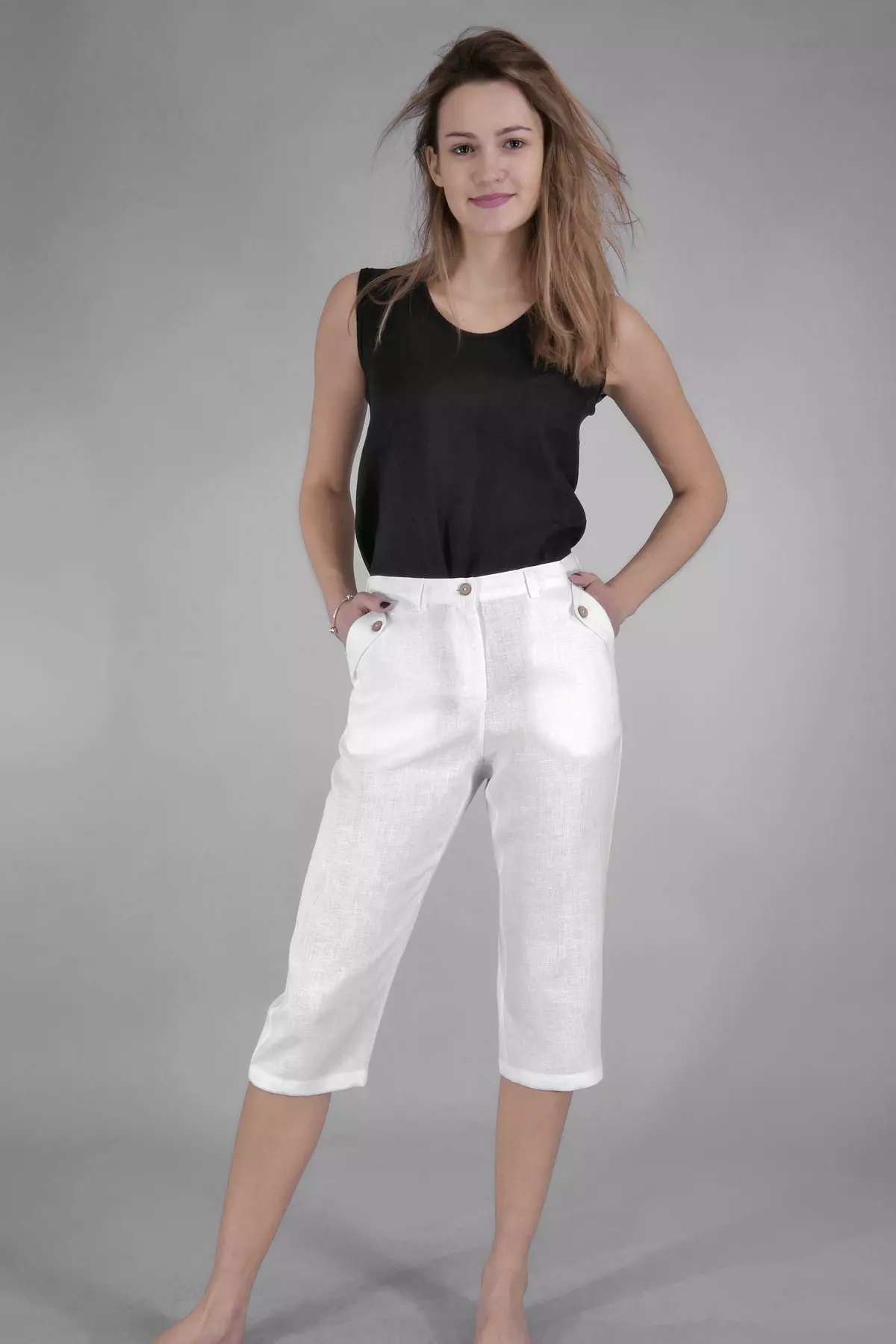 Modelka stoi w lekkim rozkroku na szarym tle. Ma na sobie białe spodnie Nata i czarną bluzkę bez rękawów. Wiatr delikatnie rozwiewa jej włosy. Spodnie mają charakterystyczne kieszenie z guzikami z przodu.