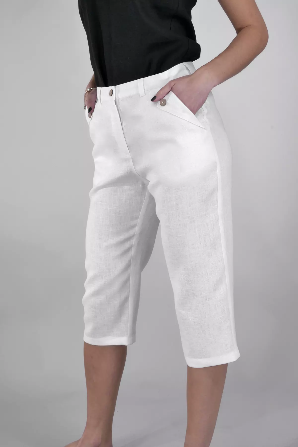 Modelka stoi w półprofilu, trzymając ręce w kieszeniach. Spodnie Nata sięgają za kolano (długość 3/4). Widać dopasowanie w biodrach i prostą nogawkę. Biały len wygląda na przewiewny.