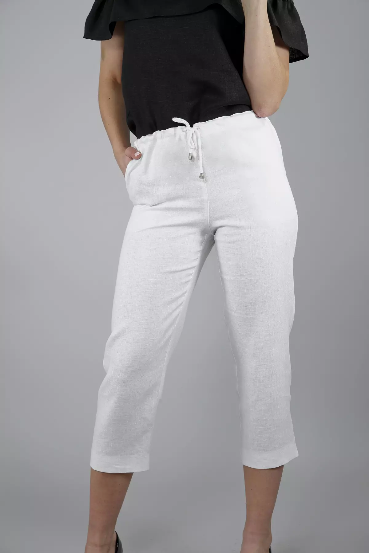 Modelka stoi w półprofilu na szarym tle. Prezentuje białe spodnie lniane Mira o długości sięgającej połowy łydki. Spodnie mają gumkę i sznurek w pasie. Zestawione są z kontrastową, czarną bluzką.