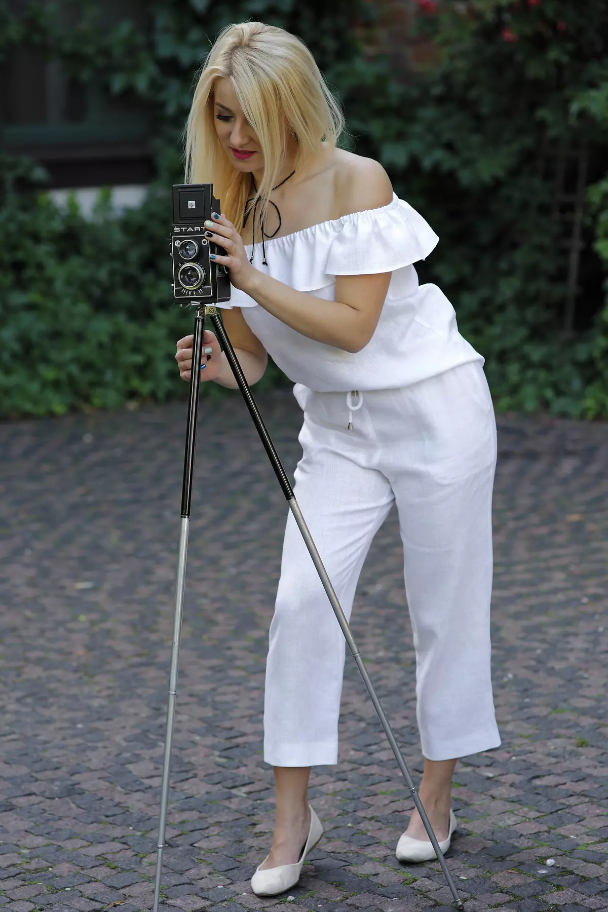 Blondynka stoi w lekkim rozkroku, pochylona nad starym aparatem fotograficznym na trójnogu. Ma na sobie biały, lniany komplet: bluzkę hiszpankę i spodnie Mira o długości 7/8. Stylizacja jest letnia i artystyczna.